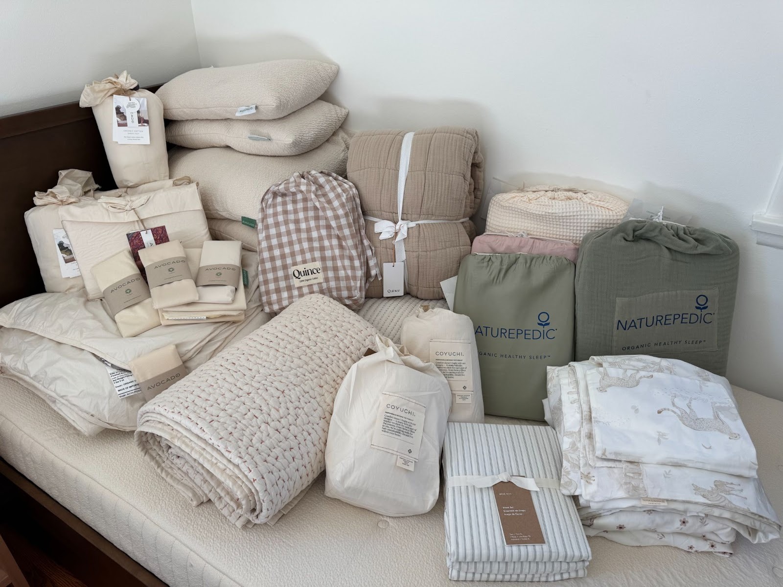 A variety of neatly stacked bedding items, including pillows, quilts, comforters, sheets, and blankets, are arranged on a bed against a white wall.
