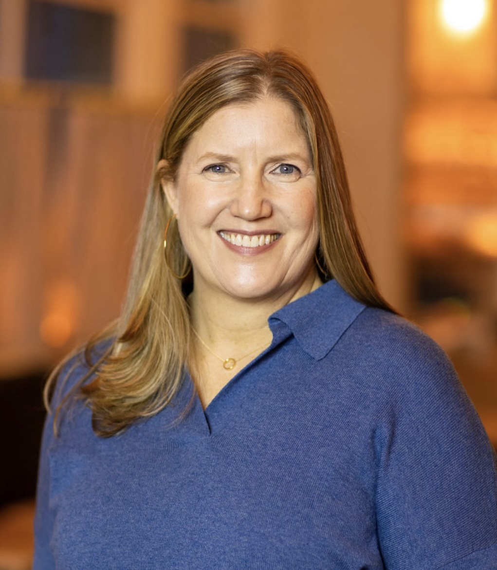 A woman with long, light brown hair is smiling at the camera. She is wearing a blue sweater and standing indoors with a blurred background.