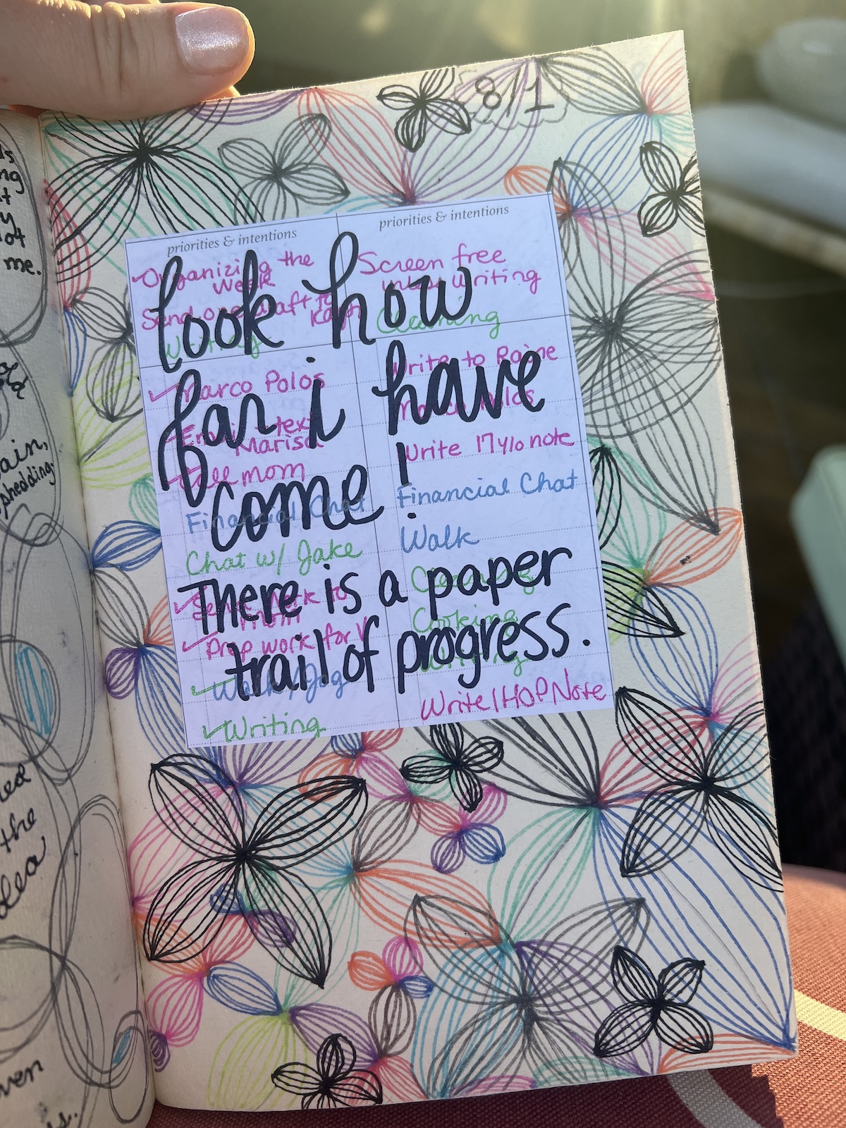 A hand holds a decorated journal page with colorful flower doodles, handwritten notes, and a quote: "Look how far I have come. There is a paper trail of progress.