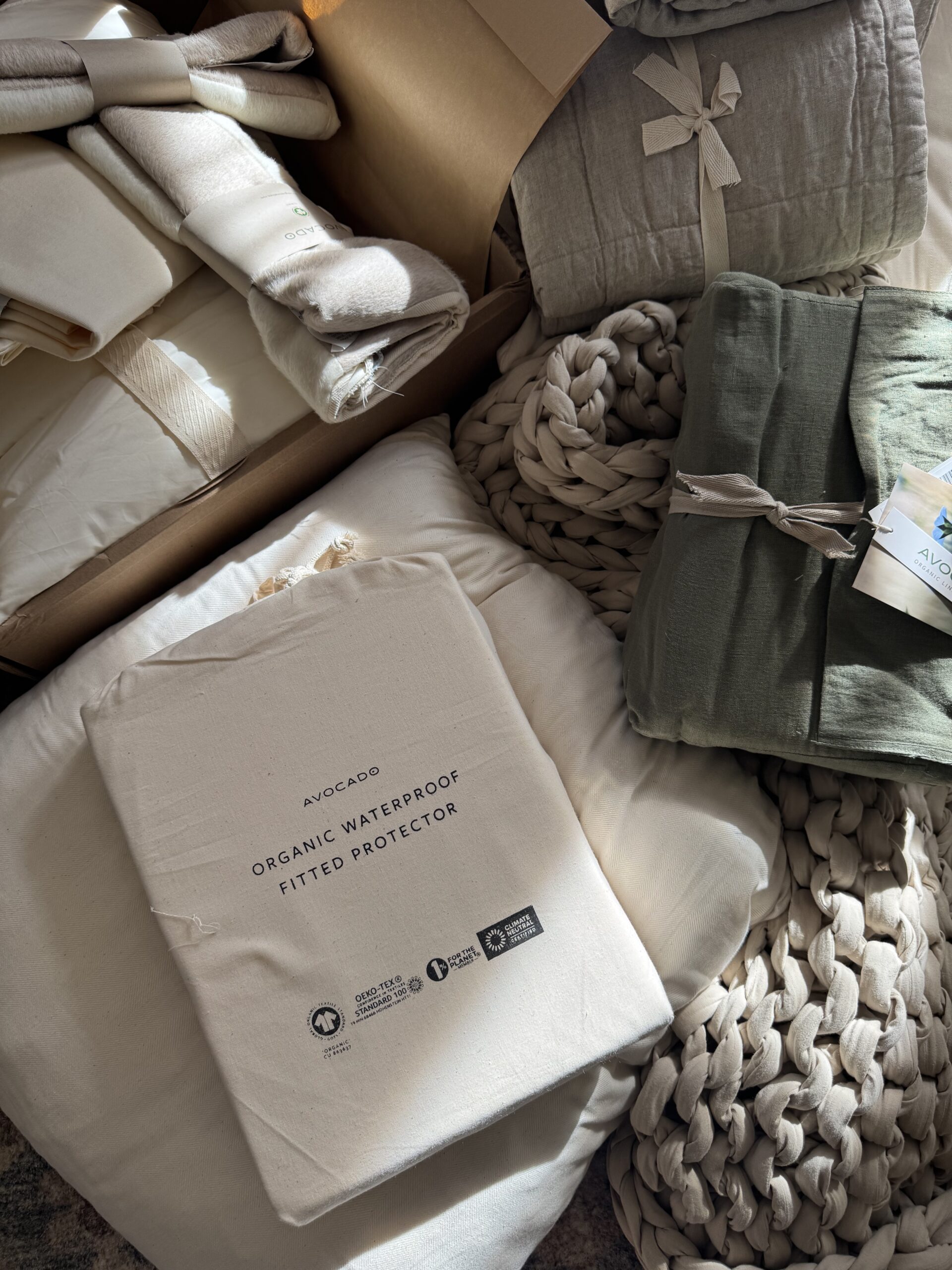 A collection of folded organic bedding items, including a waterproof fitted protector, displayed on a textured surface in soft natural light.