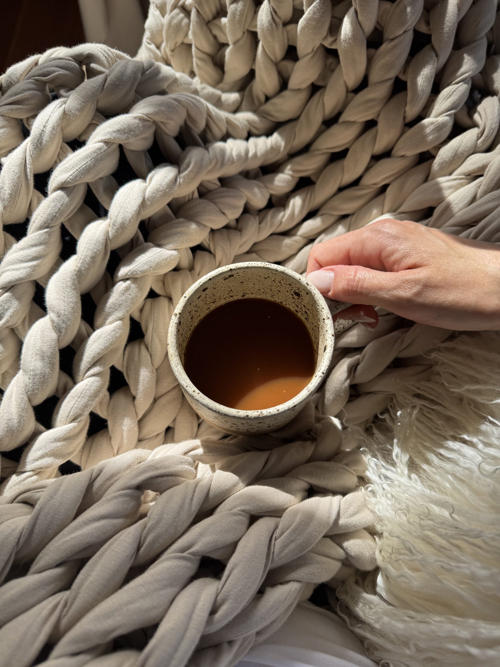 A hand holds a mug of coffee, resting on a chunky knitted blanket with sunlight casting shadows.