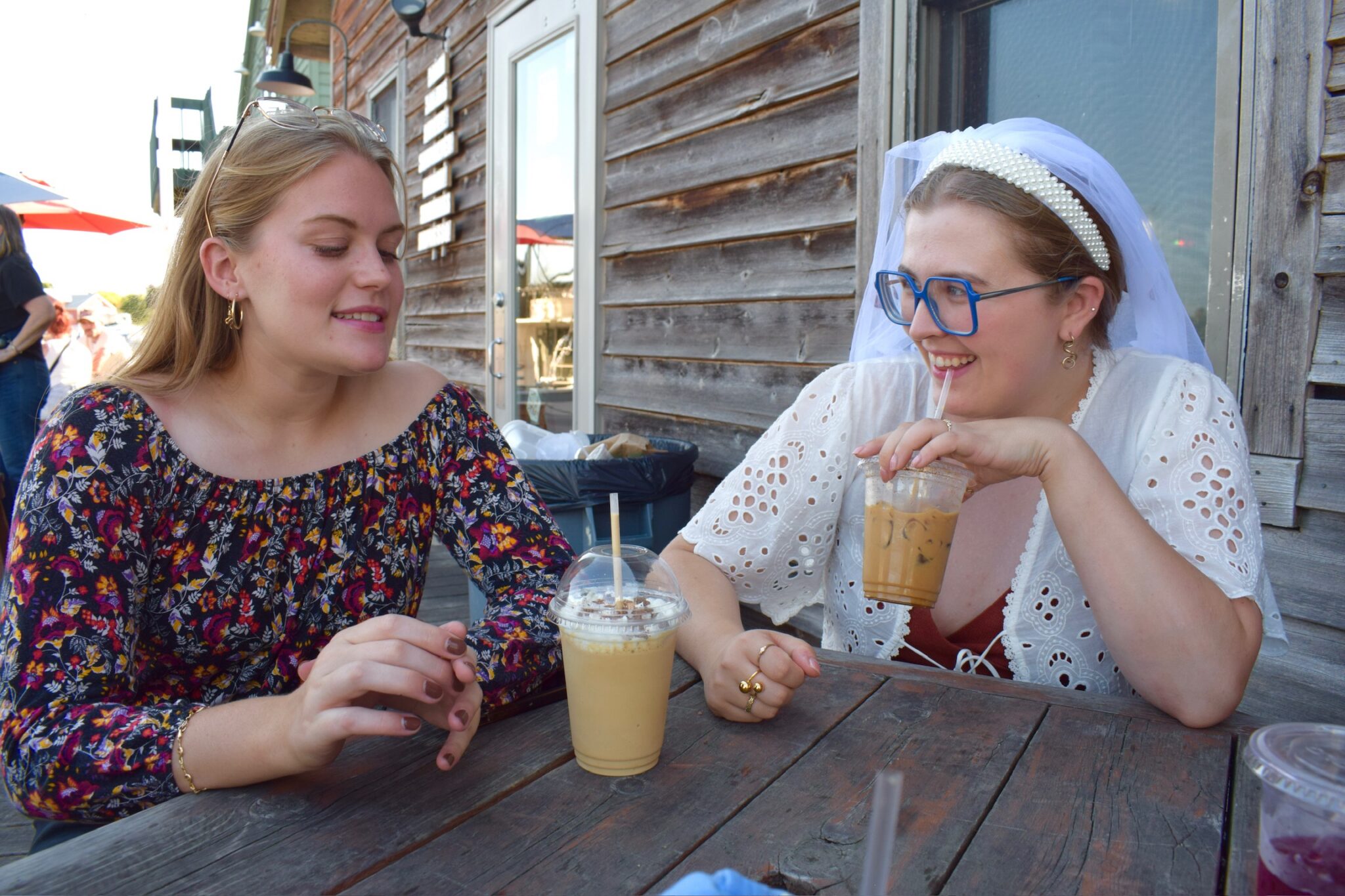Two women sit at a rustic wooden table outside, drinking iced coffee. One wears a floral dress, the other wears a white dress, blue glasses, and a veil, smiling and talking.