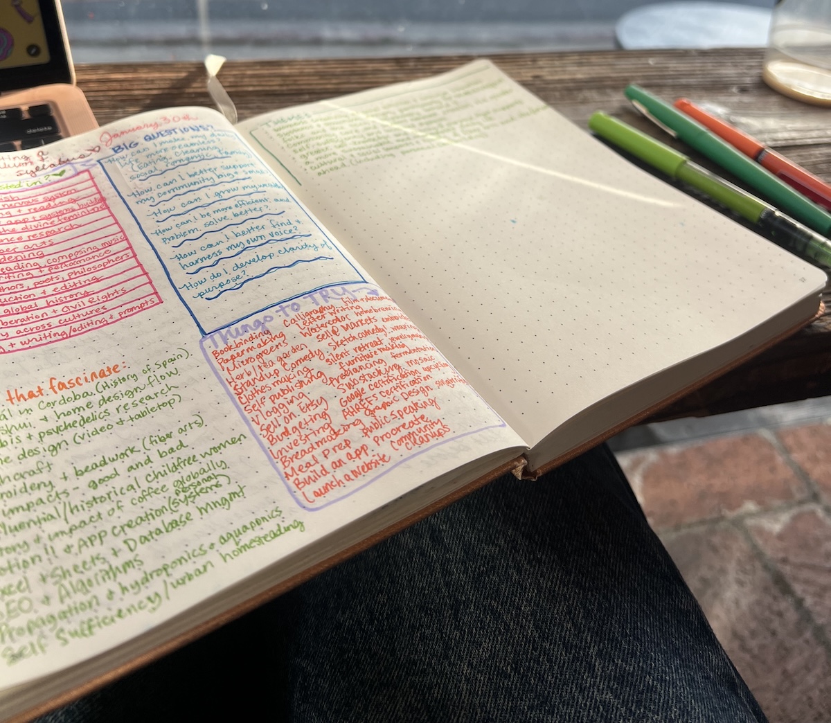 An open notebook with color-coded handwritten notes lies on someone's lap, next to several colored pens on a wooden table by a window.