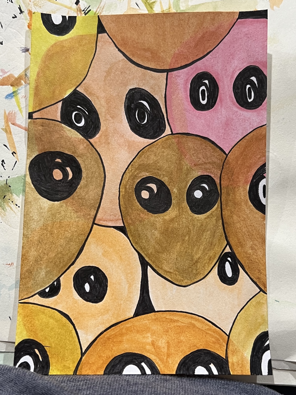 A watercolor painting shows overlapping oval shapes in earth tones with large black and white eyes, outlined in black, creating an abstract composition.
