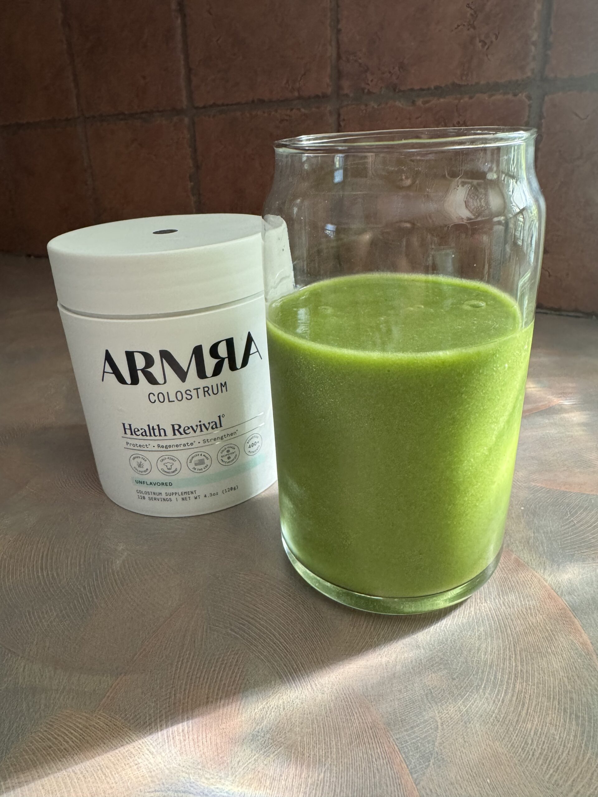 A tub of ARMRA Colostrum powder sits next to a glass filled with a bright green smoothie on a countertop.