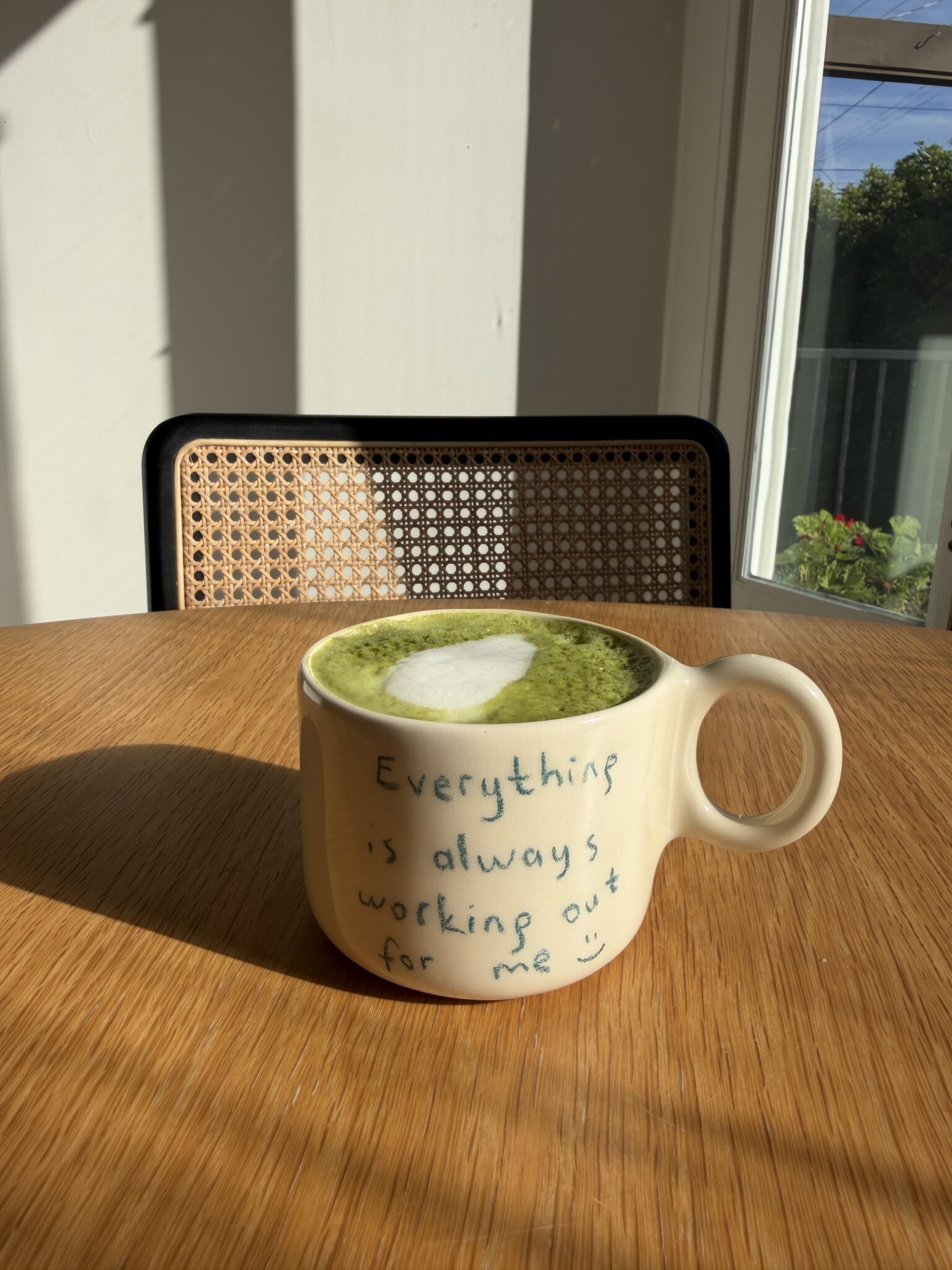 A cup of green matcha latte with heart-shaped foam sits on a wooden table. The mug reads, "Everything is always working out for me :)" A window and chair are in the background.