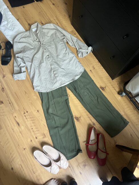 A light gray button-up shirt and green pants are laid out on a wooden floor, surrounded by three pairs of shoes: black heels, beige flats, and red Mary Janes.