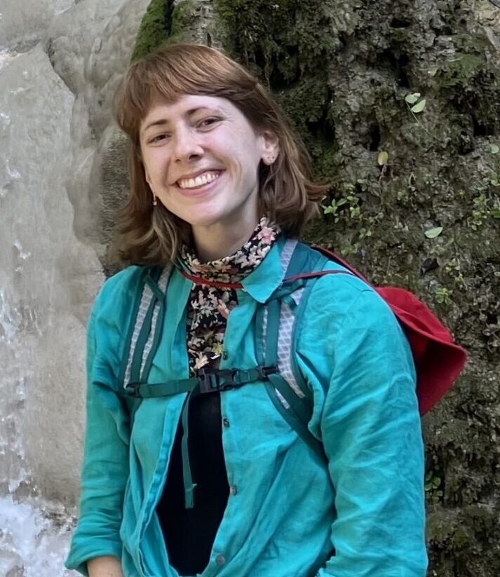 A person wearing a teal shirt, pink shorts, and a backpack stands in front of a rocky waterfall, smiling at the camera.