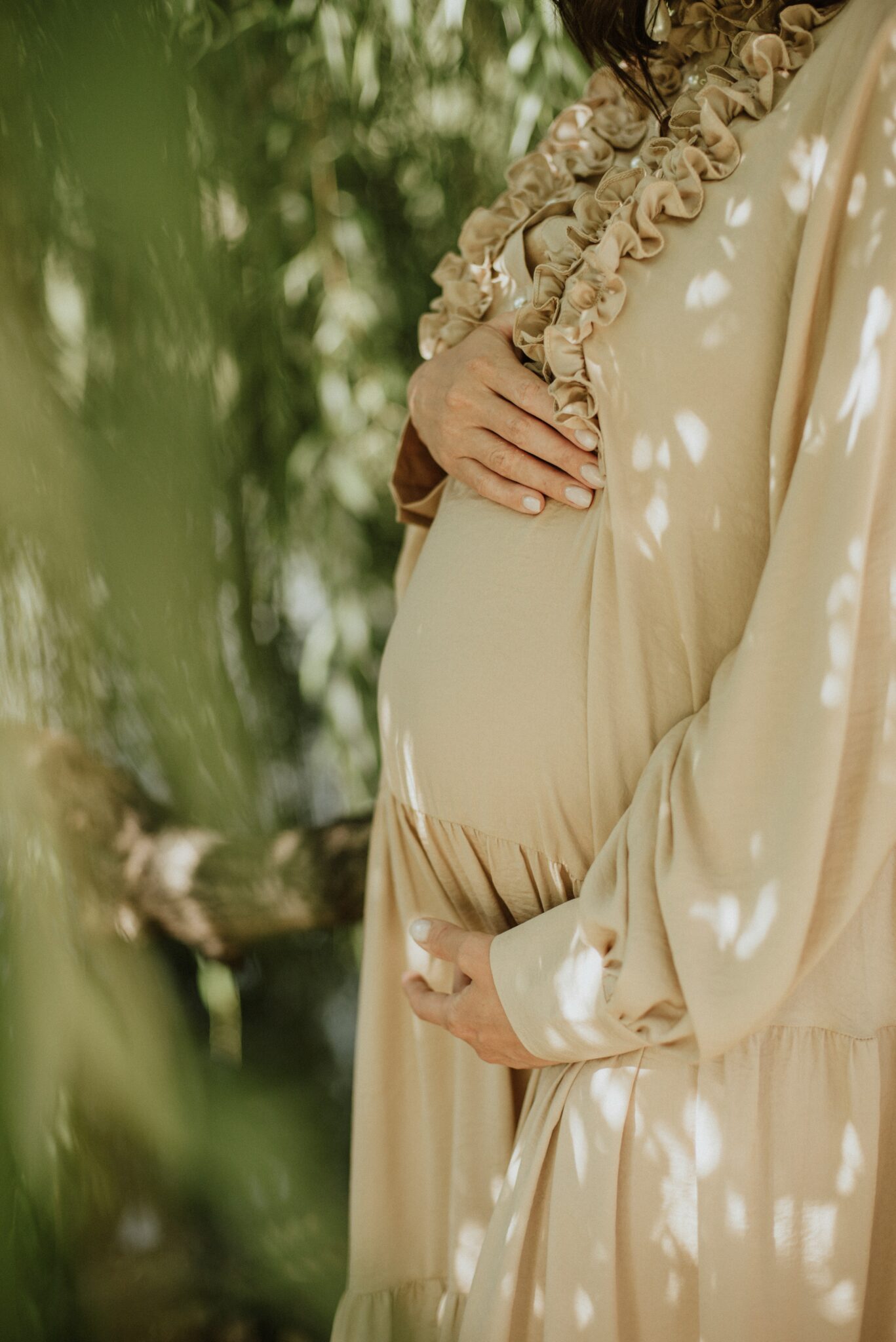 A person wearing a beige dress is standing outdoors, holding their pregnant belly with both hands.