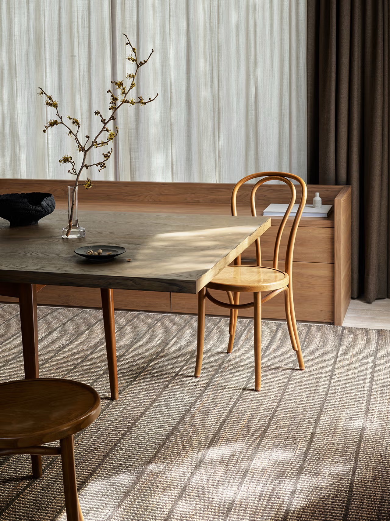 A wooden dining table with two chairs, a glass vase with branches, a black bowl, and a small dish on a patterned rug in a sunlit room with brown curtains and a wooden bench.