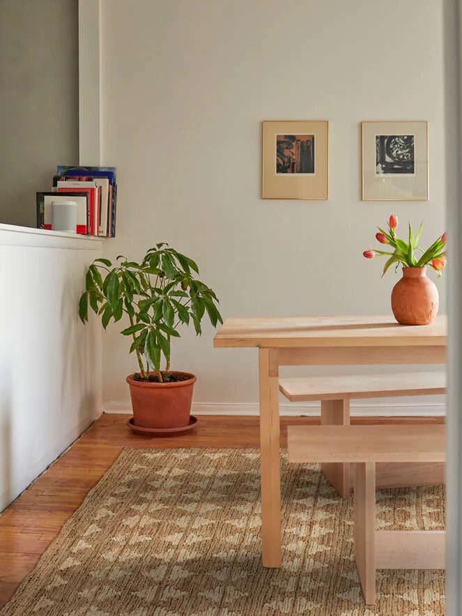 A minimal dining area with a light wood table and bench, a potted plant, a vase of flowers, framed art on white walls, and a patterned rug on a wooden floor.