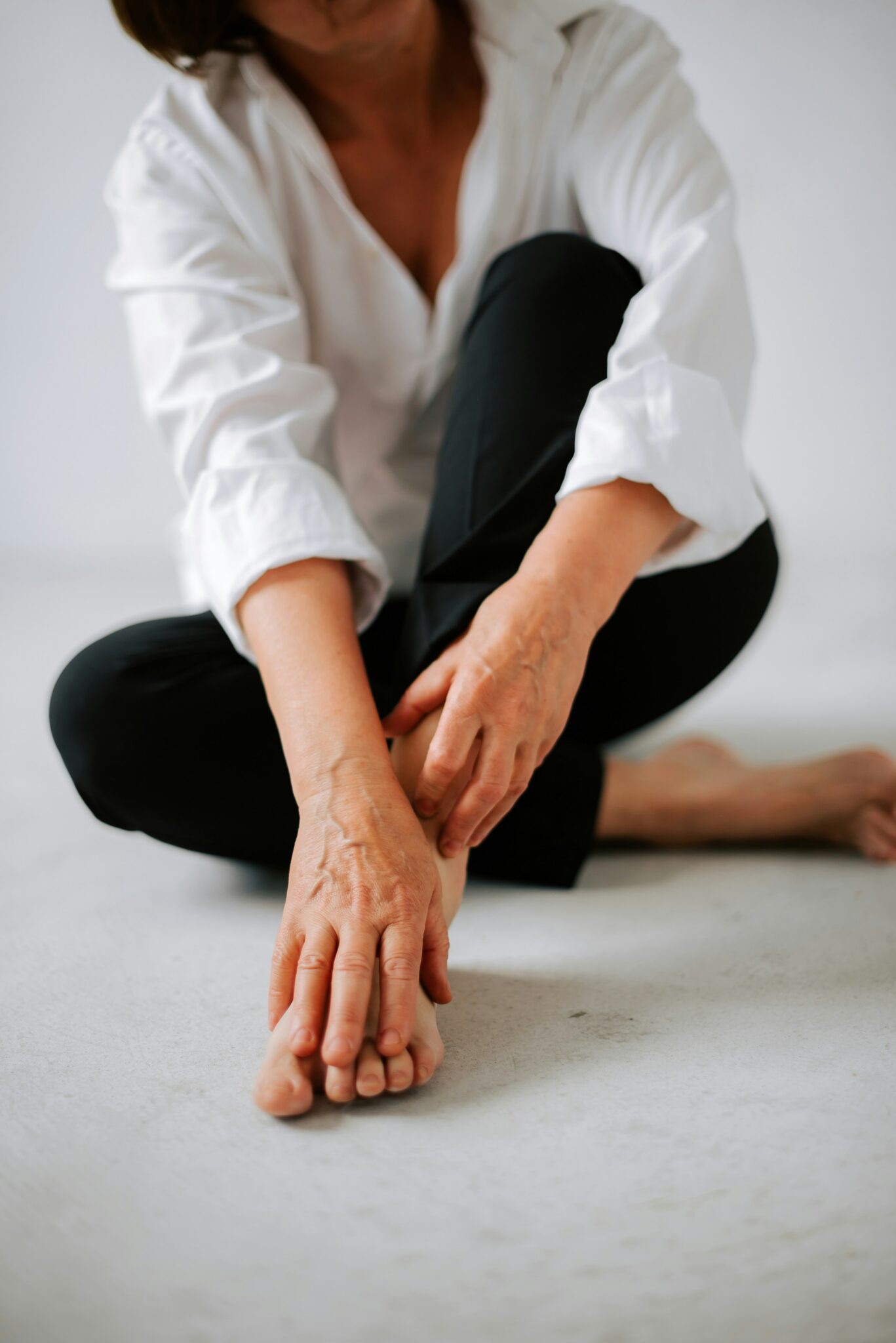 A person in a white shirt and black pants sits on the floor touching their foot and ankle with their hands.