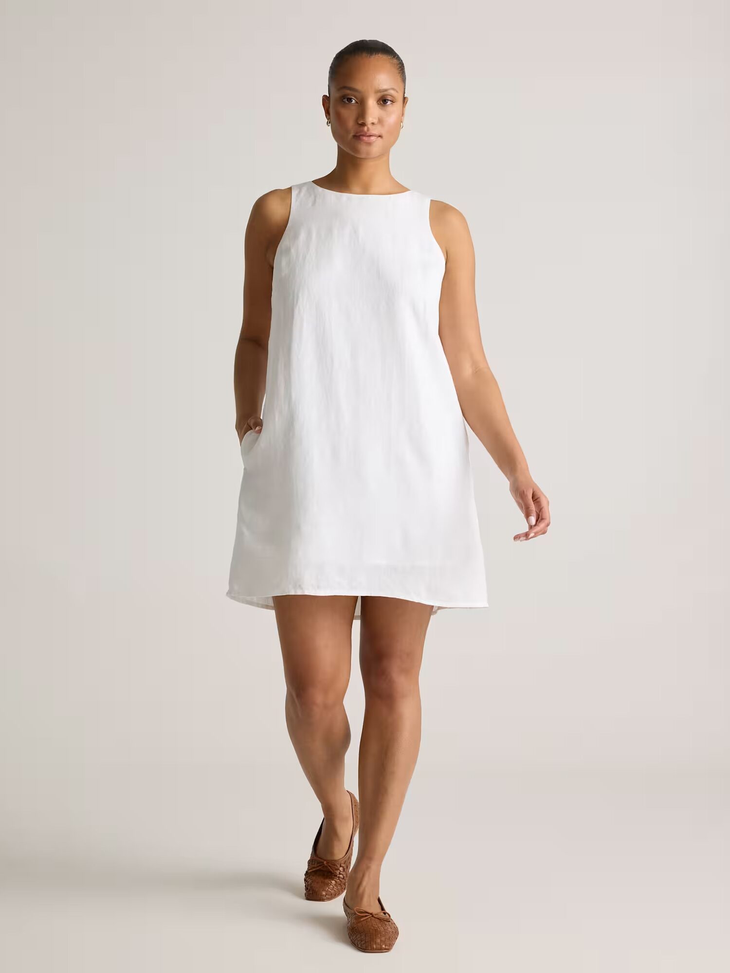A woman stands facing forward wearing a sleeveless, above-the-knee white dress and brown woven flats against a plain background.