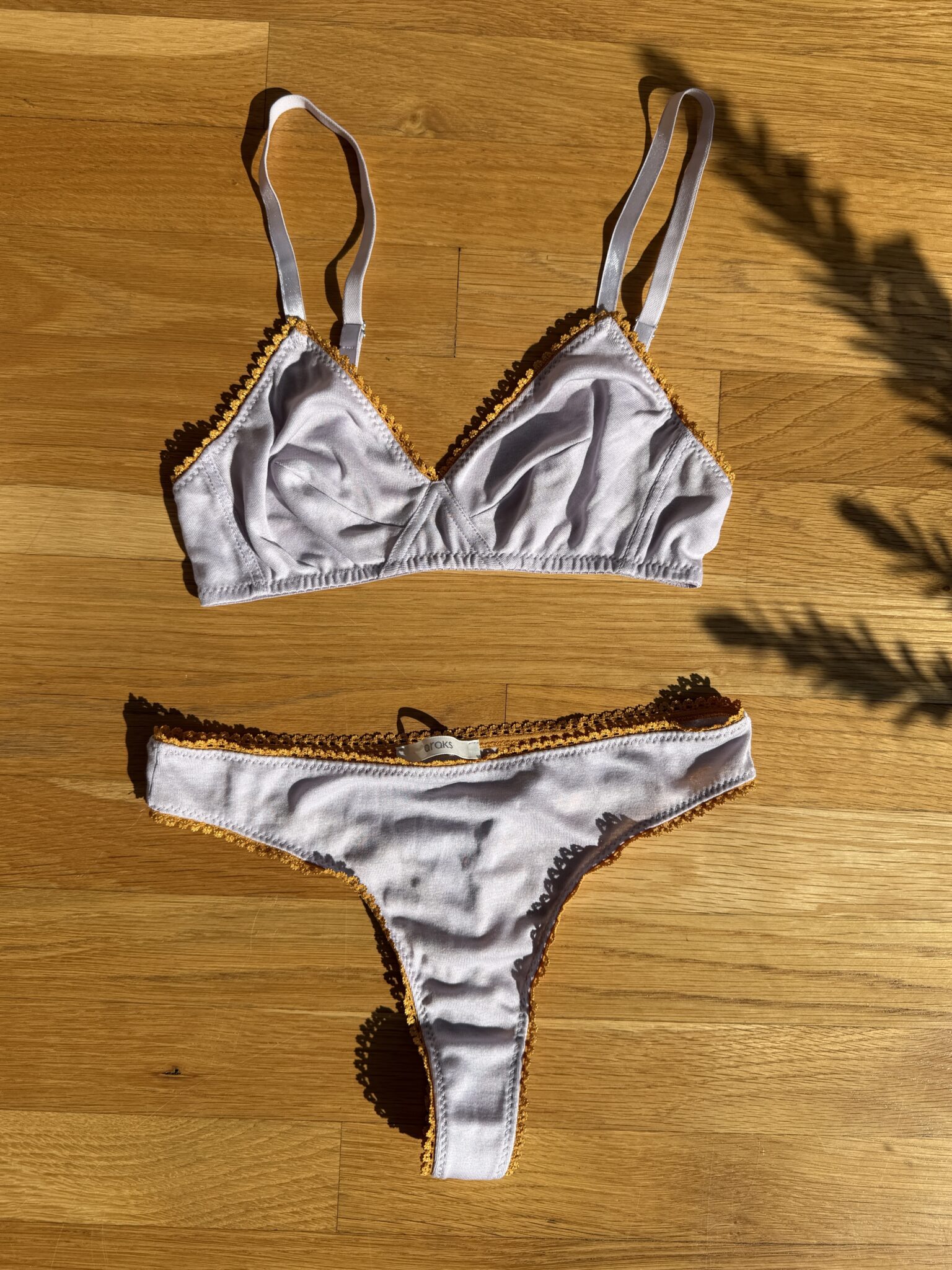 A lilac bralette and matching thong with orange trim are laid flat on a wooden floor in direct sunlight.
