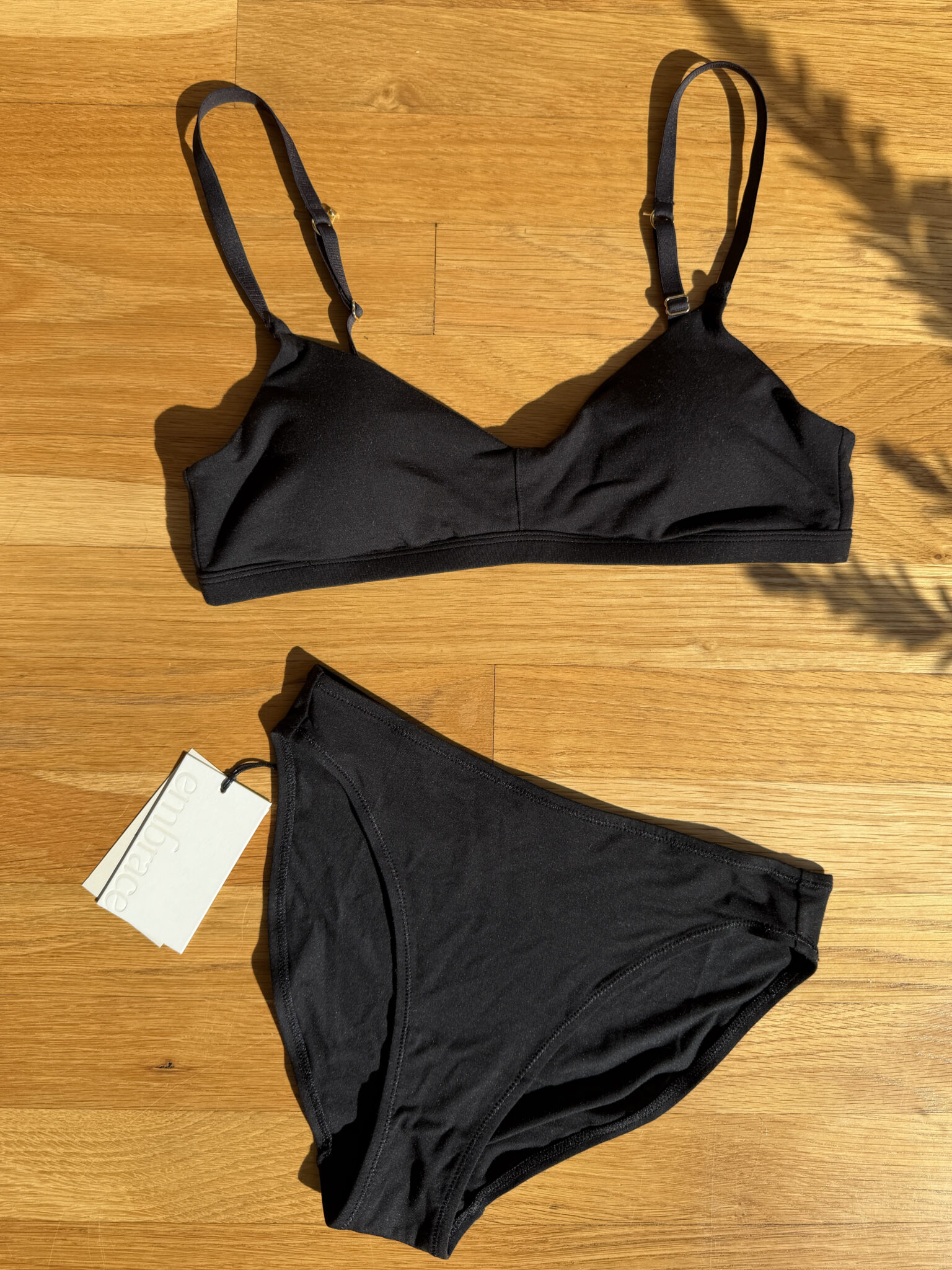 A black bralette and matching black bikini briefs are laid flat on a wooden floor, with a product tag attached to the briefs.