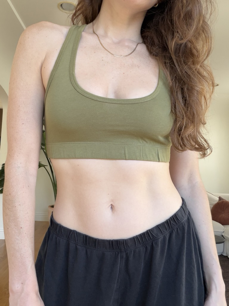 A person wearing an olive green sports bra and black pants stands indoors, partially visible from the neck down.