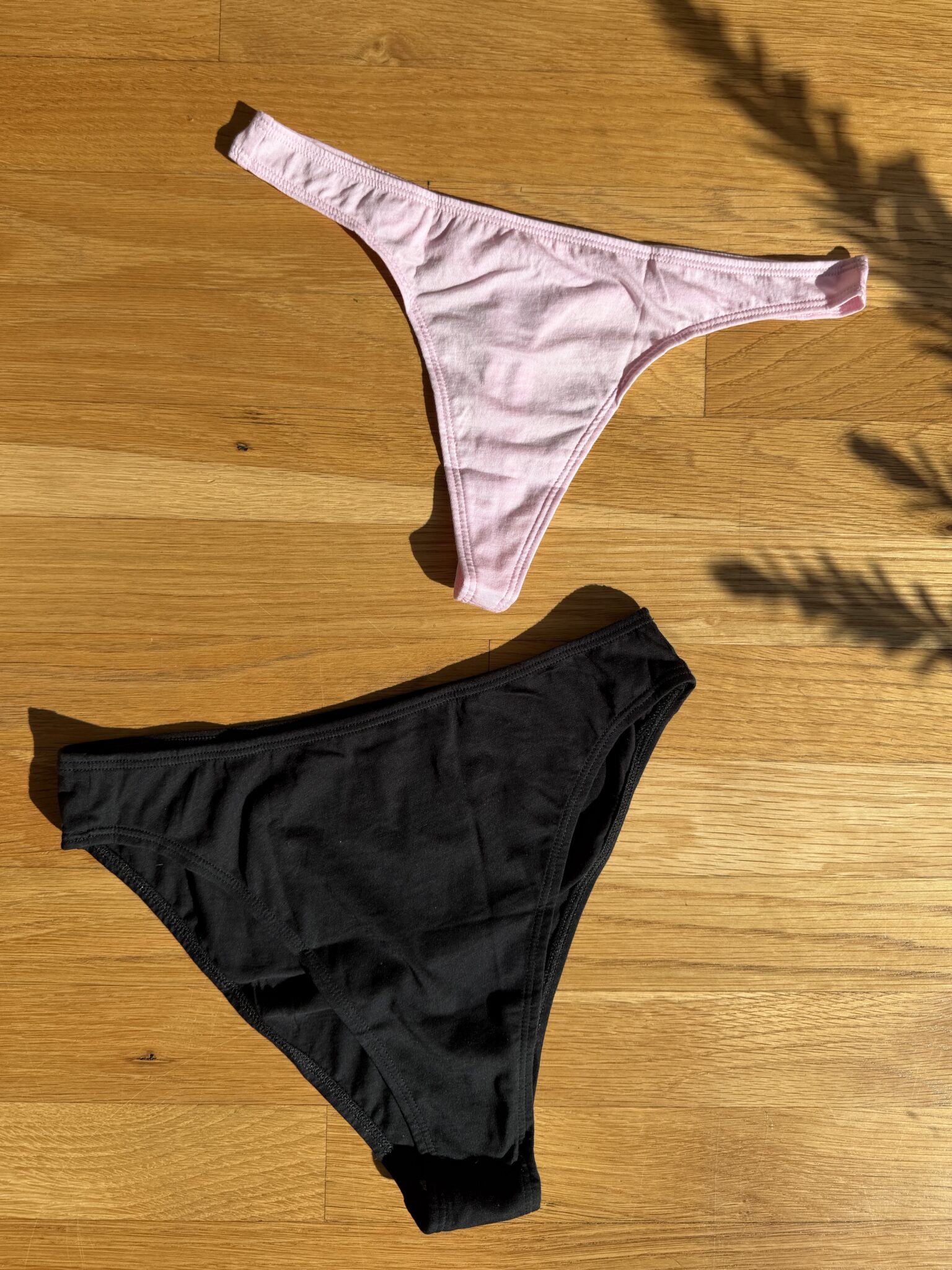 A pink thong and a pair of black bikini-style underwear are laid out on a wooden floor in sunlight.