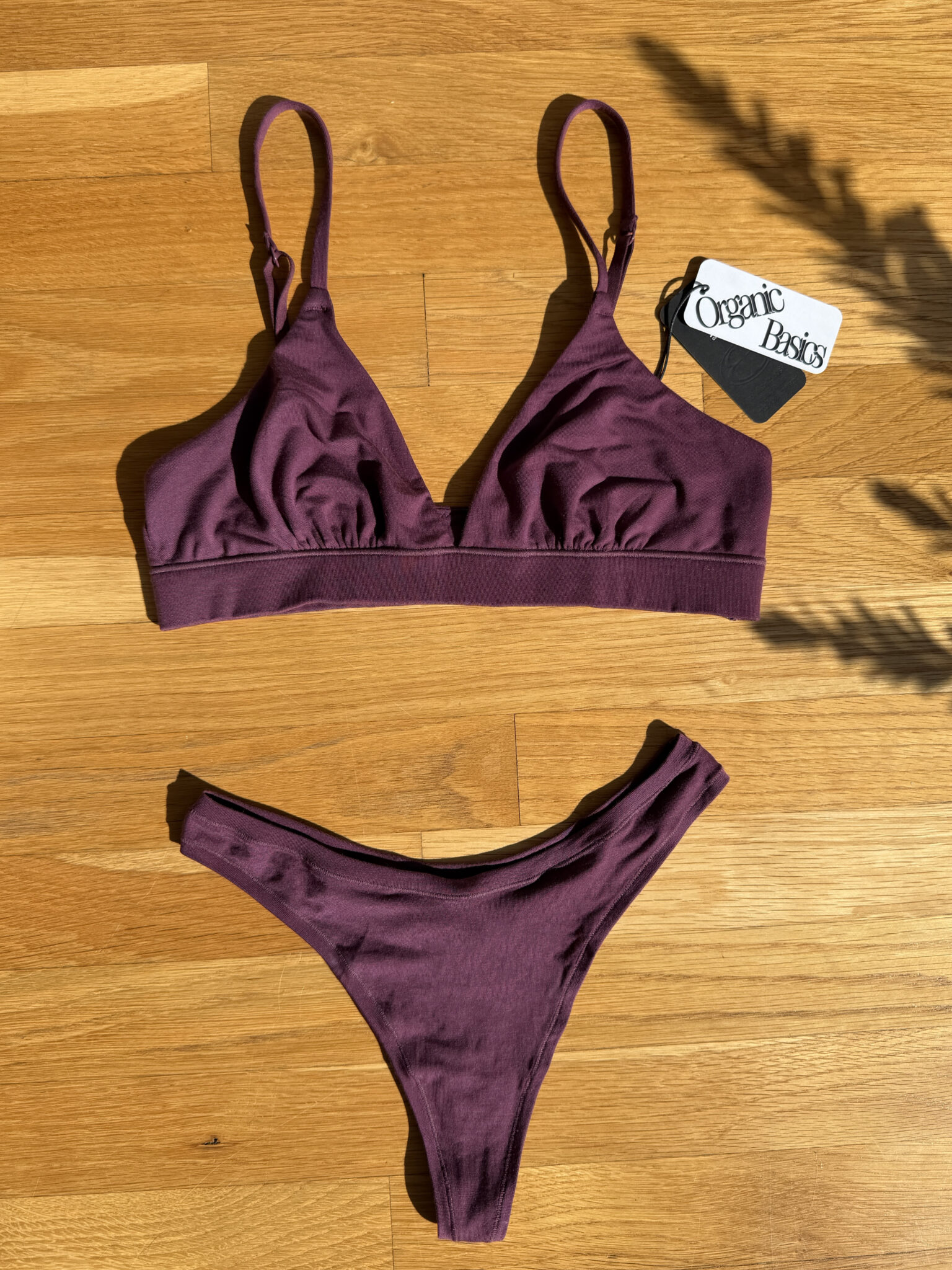 A purple bralette and matching thong underwear set laid flat on a wooden floor, with an Organic Basics tag placed nearby.