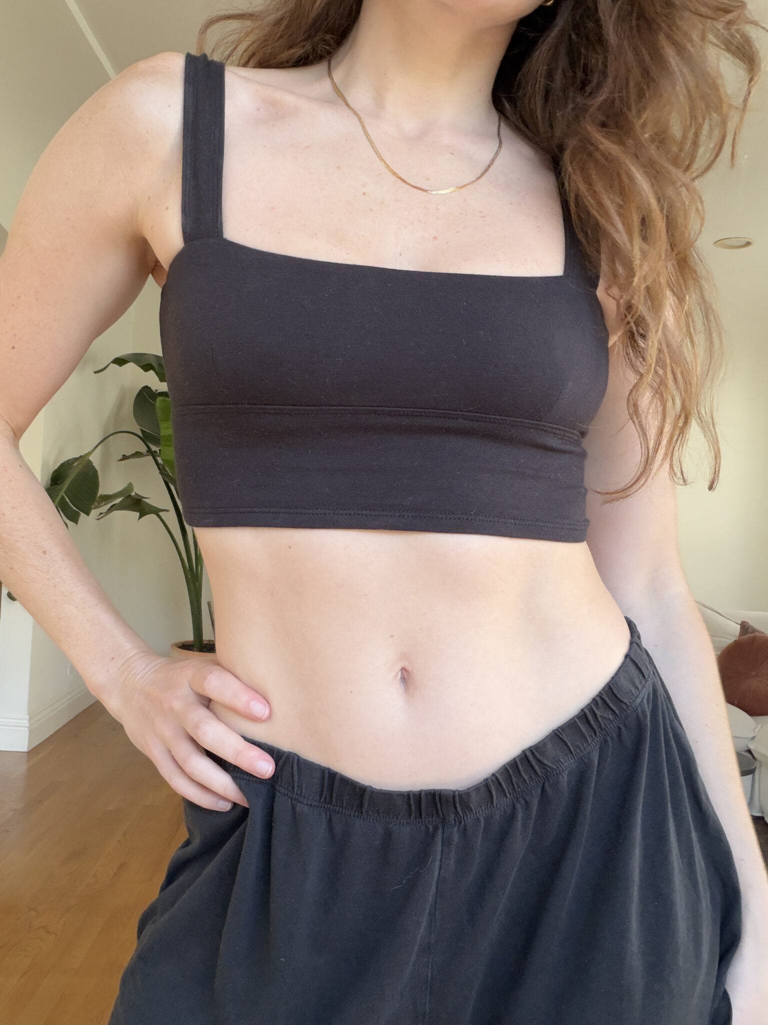 A woman wearing a black crop top and black pants stands indoors with one hand on her hip.