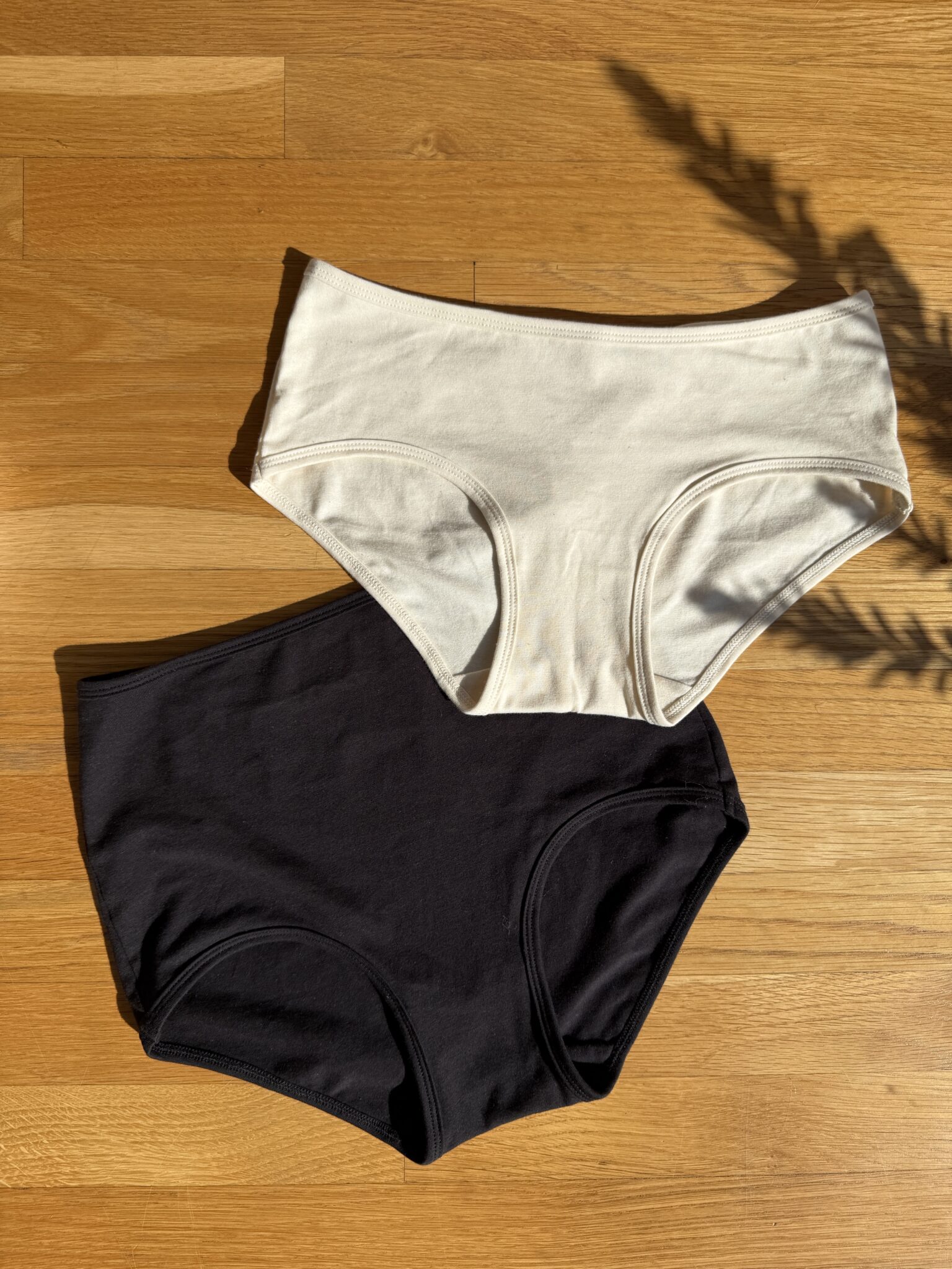 Two pairs of women's underwear, one beige and one black, are laid flat on a wooden floor in sunlight.