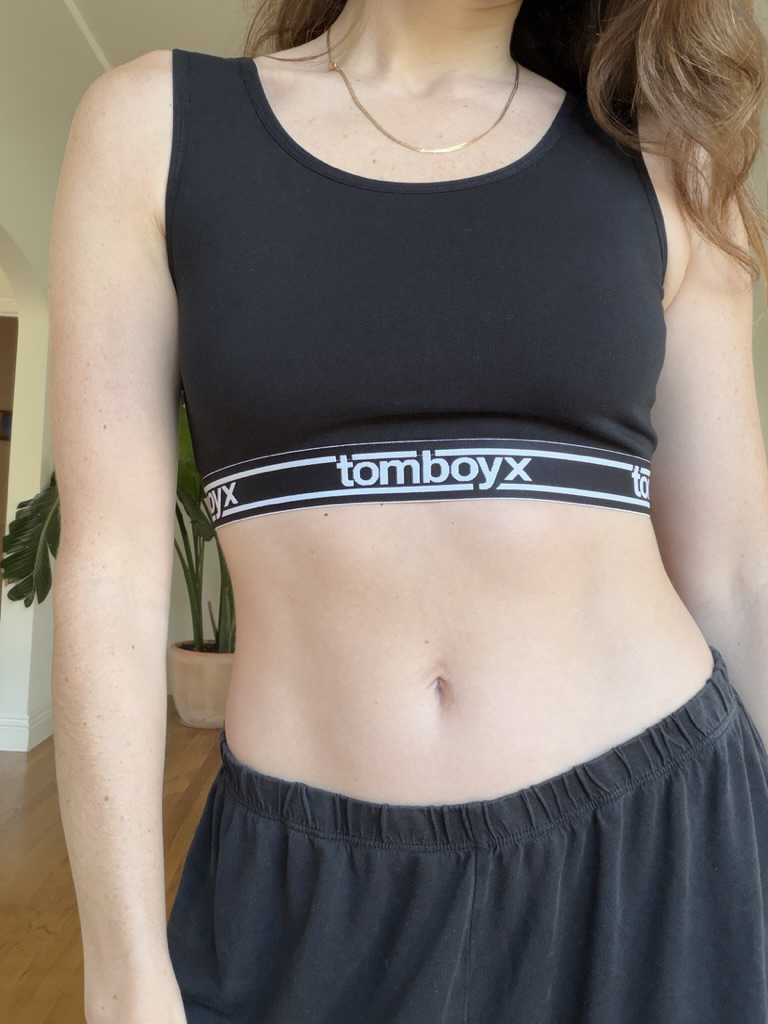 Person wearing a black crop top with "tomboyx" branded elastic band and black pants, standing indoors next to a large plant.