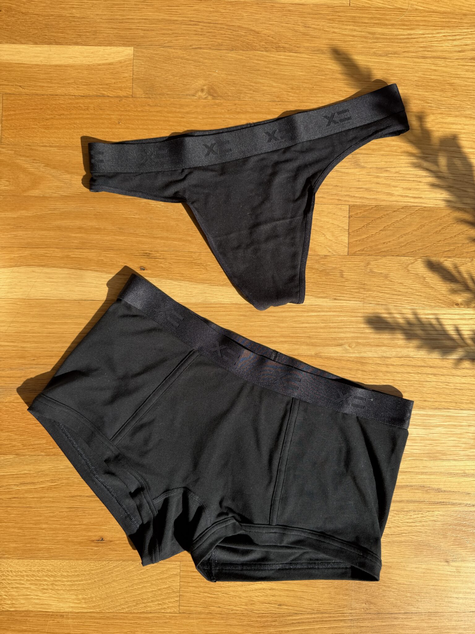 A black thong and a pair of black boxer briefs are laid out on a wooden surface in natural sunlight.
