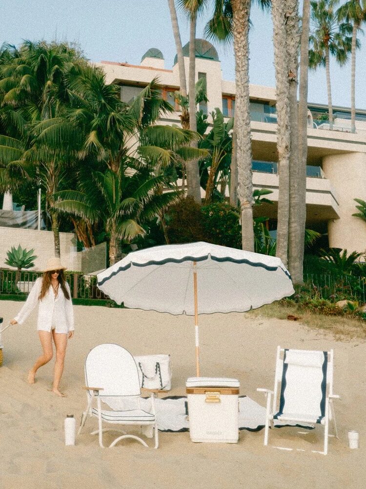 Two people on a sandy beach setting up with a large umbrella, cooler, and chairs. One is pulling a cart while the other arranges items. Palm trees and buildings are in the background.