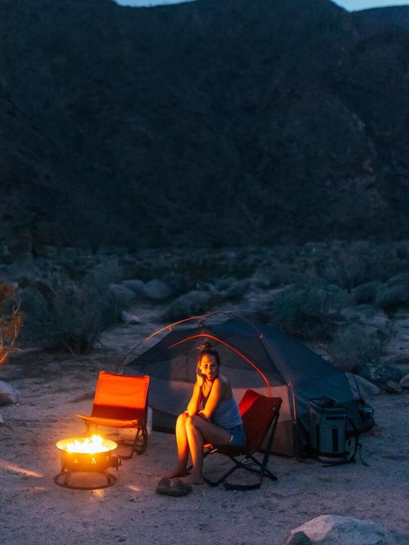 Person sitting on a chair beside a campfire, near a tent, in a rocky, desert-like area at dusk.