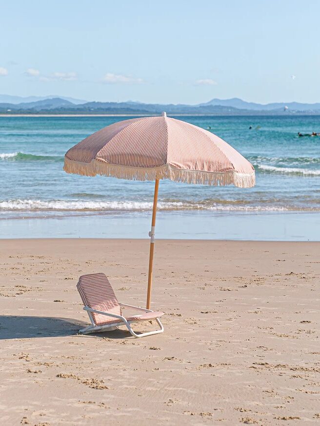 A striped beach umbrella and lounge chair are set up on a sandy beach with calm ocean waves and distant mountains in the background.