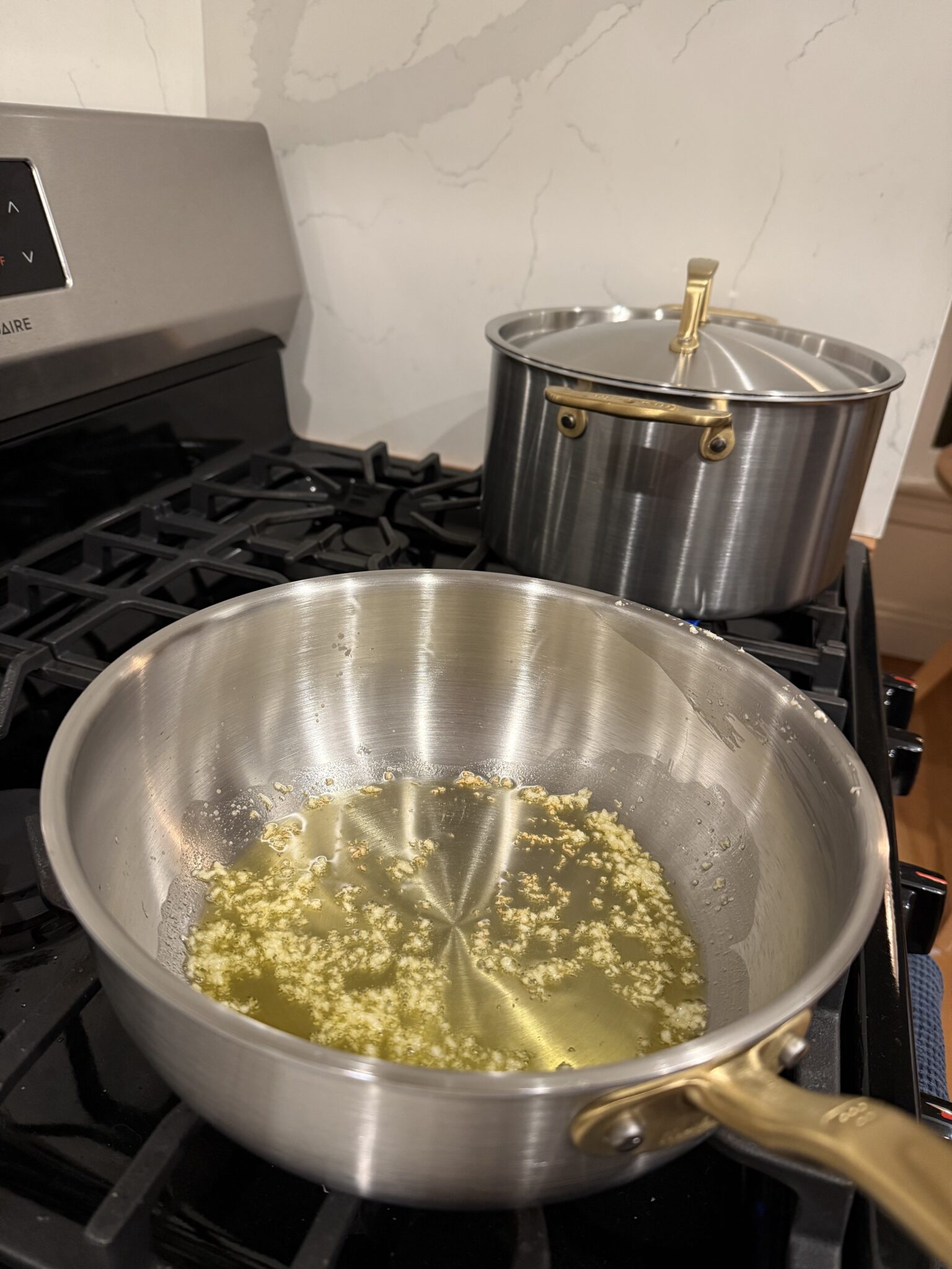Minced garlic sautéing in oil in a stainless steel saucepan on a stove, with another lidded pot in the background.