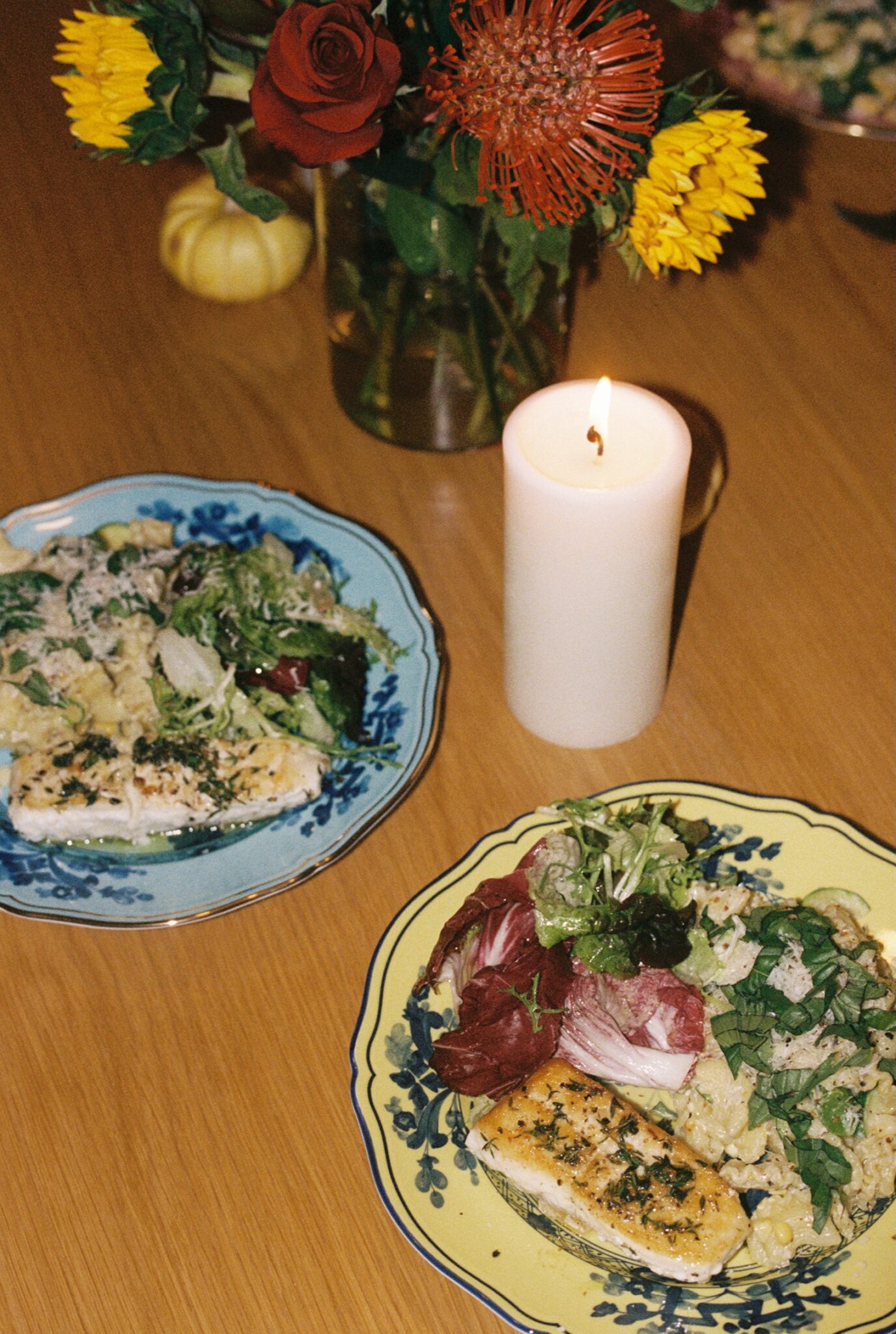 Two plates of salad and grilled fish sit on a wooden table beside a lit white candle and a vase of assorted flowers.
