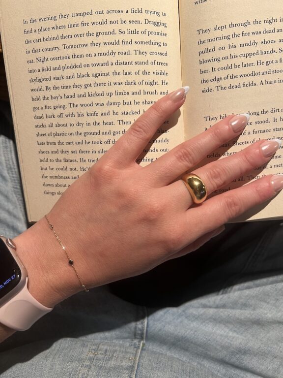 A hand with manicured nails, a gold ring, and a bracelet rests on an open book. The person is wearing a smartwatch with a light pink band.