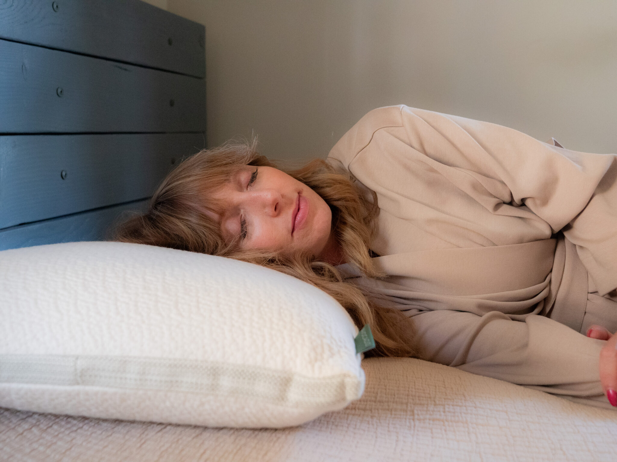 A woman with long hair is lying on her side on a bed, resting her head on a white pillow and wearing a beige robe.