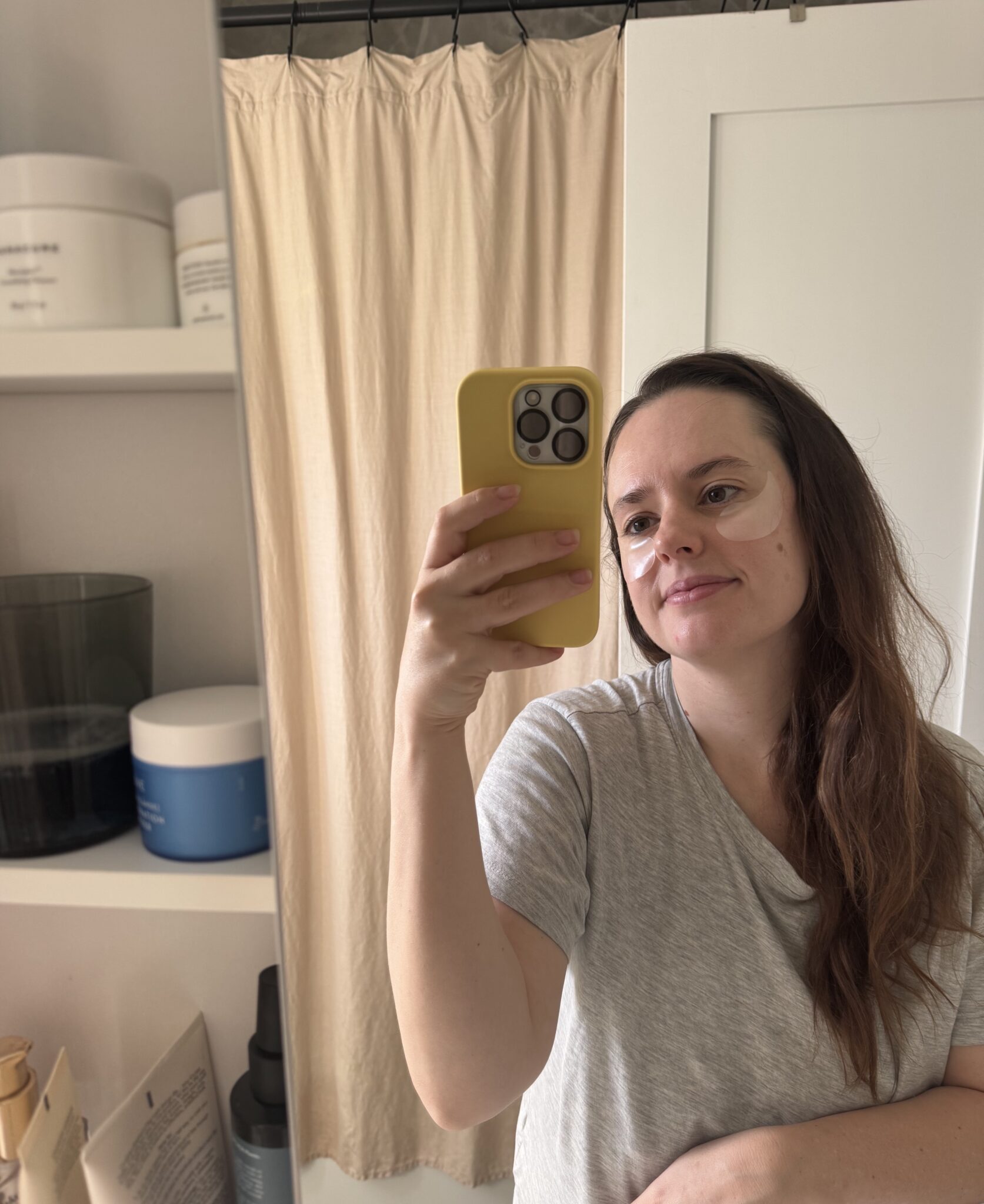 A woman with under-eye patches takes a selfie in front of a mirror in a bathroom, holding a yellow phone and wearing a grey t-shirt.