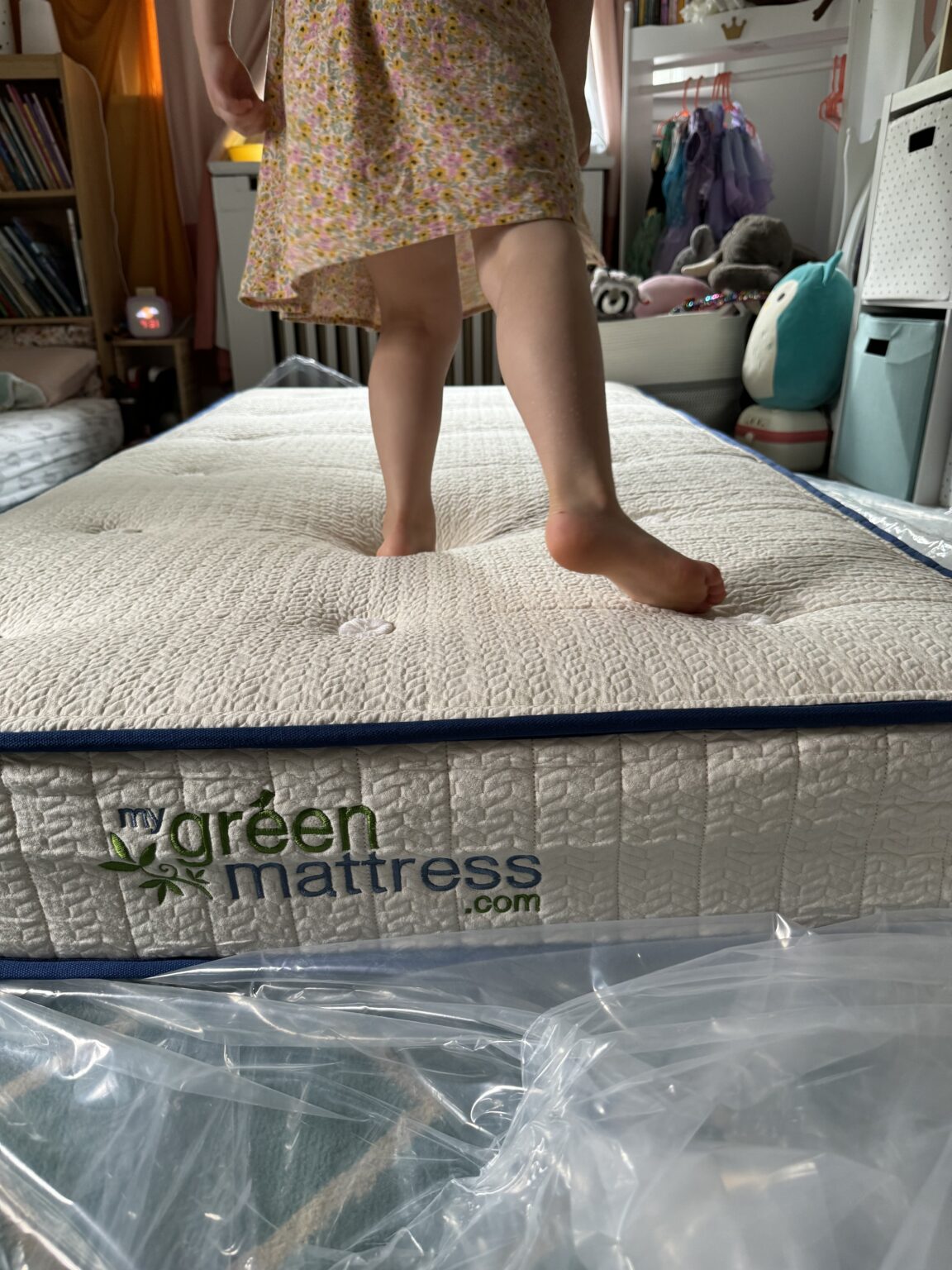 A child in a floral dress stands on an organic mattress in a bedroom, with sunlight streaming through the window and plastic packaging visible in the foreground.