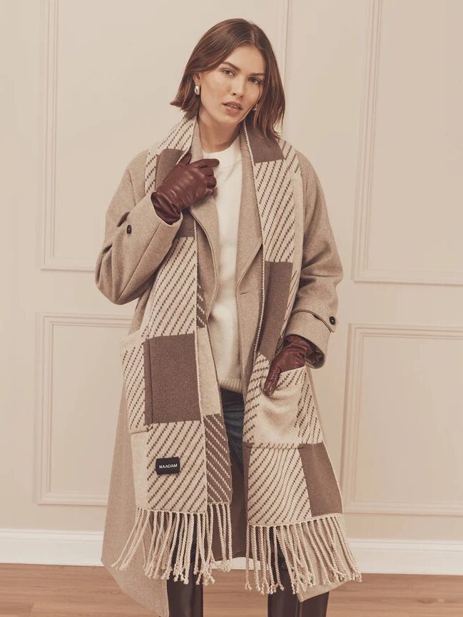 A woman wearing a long beige coat, matching patterned scarf, brown leather gloves, and white top stands in front of a cream wall with panel detailing.