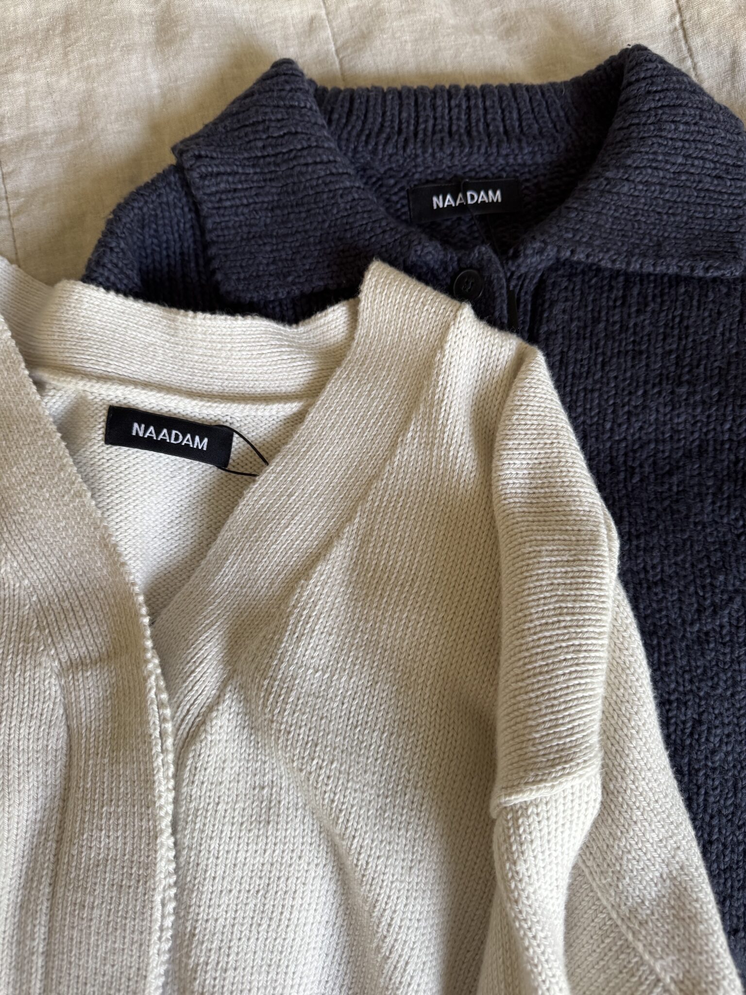 Two knit cardigans, one cream and one dark navy, both with NAADAM labels, are laid flat on a beige fabric surface.