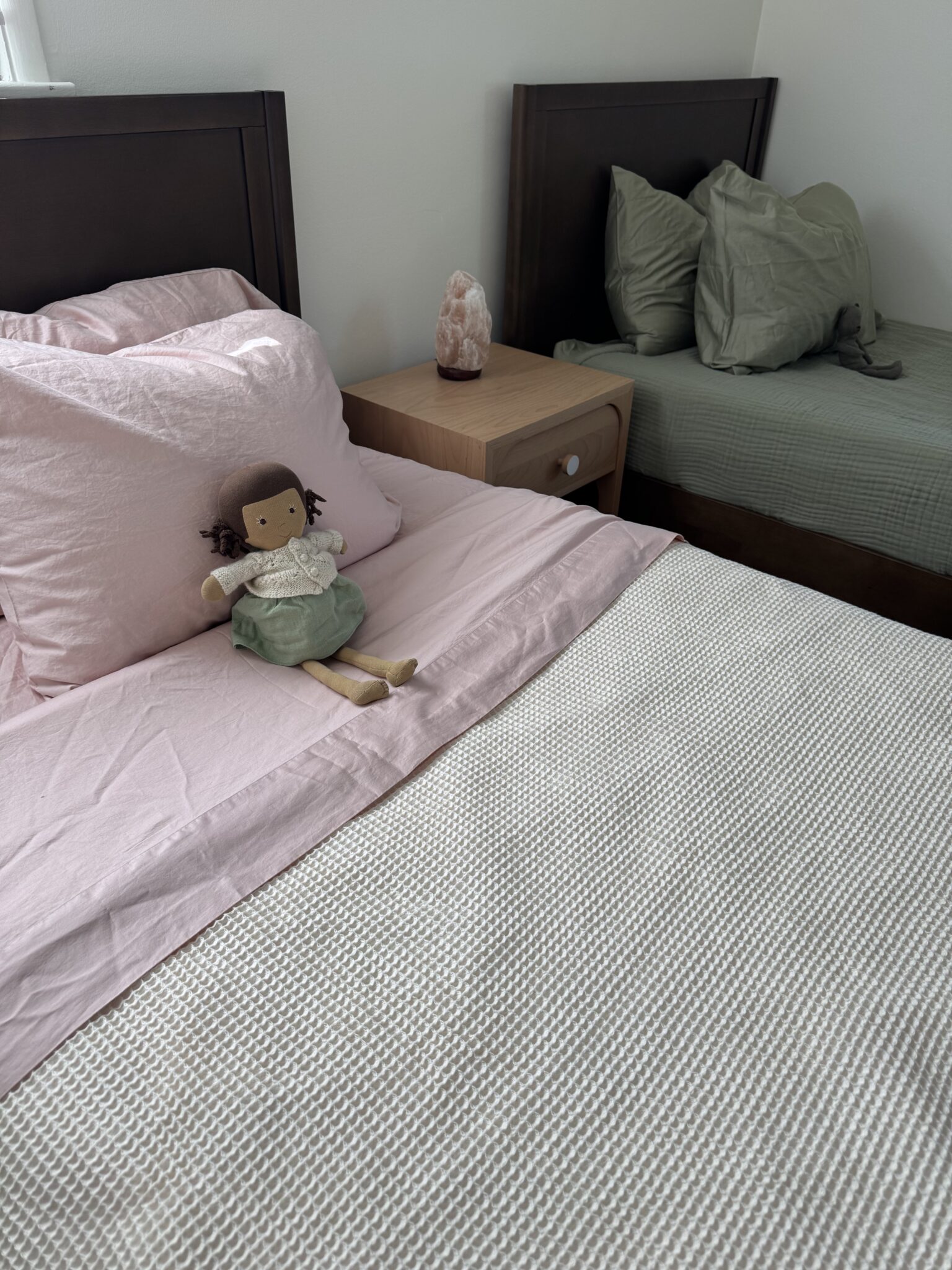 Two neatly made beds with pastel bedding, a nightstand between them, and a cloth doll placed on the bed with pink sheets.
