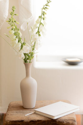 A beige vase with white flowers sits on a wooden table next to a closed white notebook and pen, with a small dish in the background.