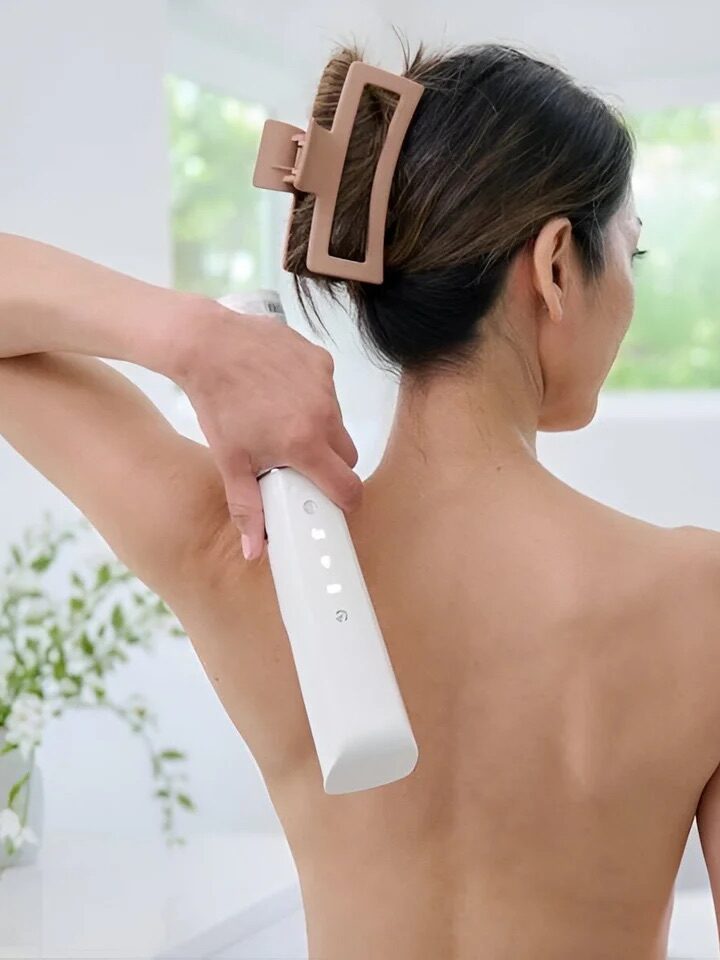 A woman with a hair clip uses a handheld device on her back in a bright room with a vase of flowers in the background.