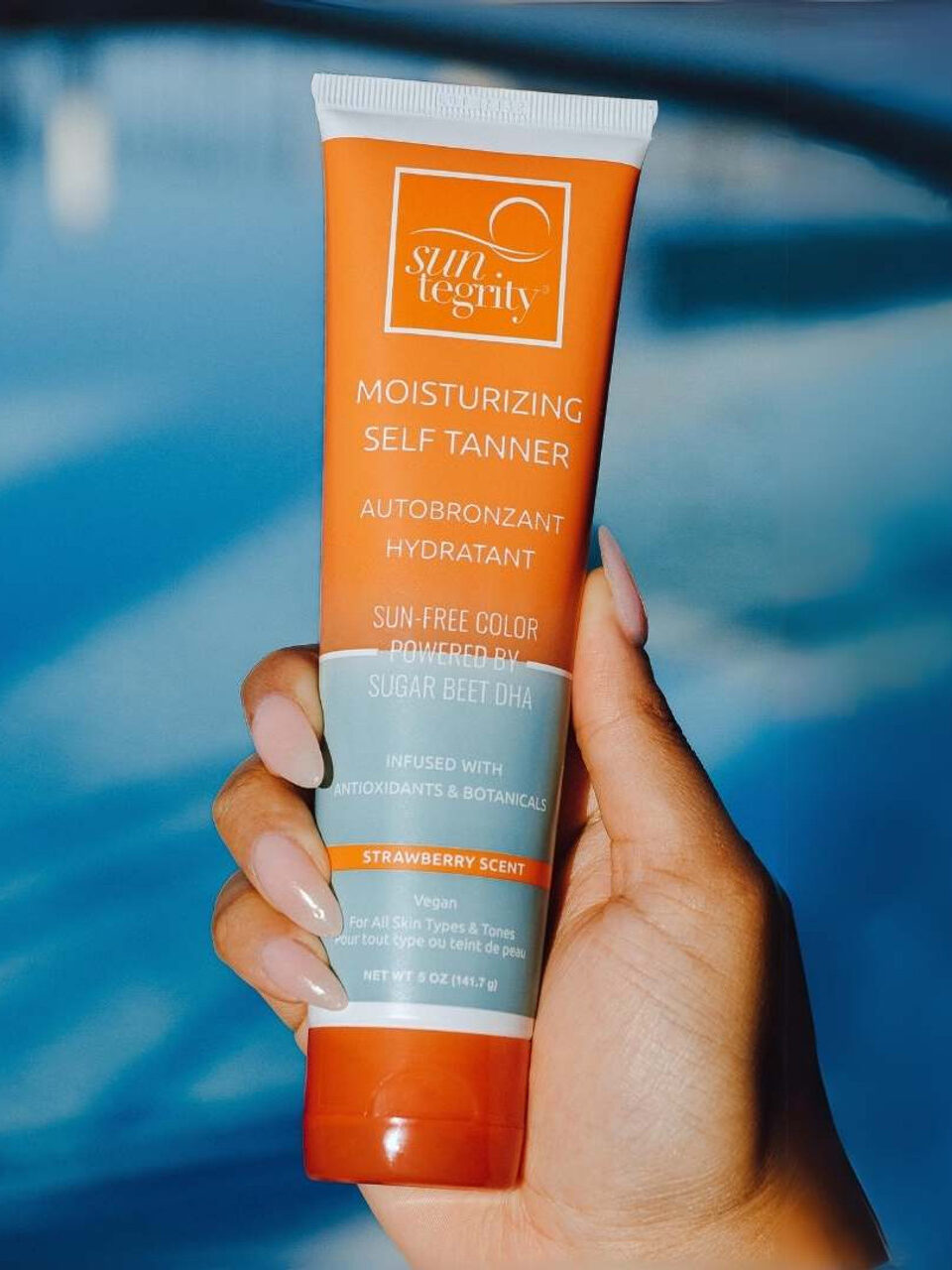 A person holding a tube of Suntegrity Moisturizing Self Tanner in front of a blue, out-of-focus background.