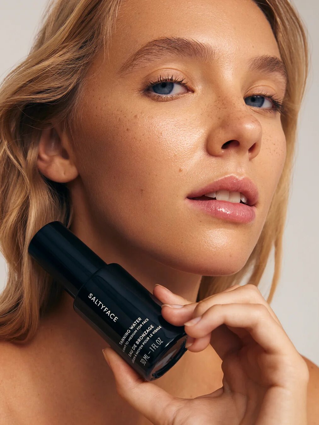 A woman with light skin and blonde hair holds a black bottle labeled "SALTYFACE" near her face, showcasing dewy, natural makeup and clear skin against a neutral background.