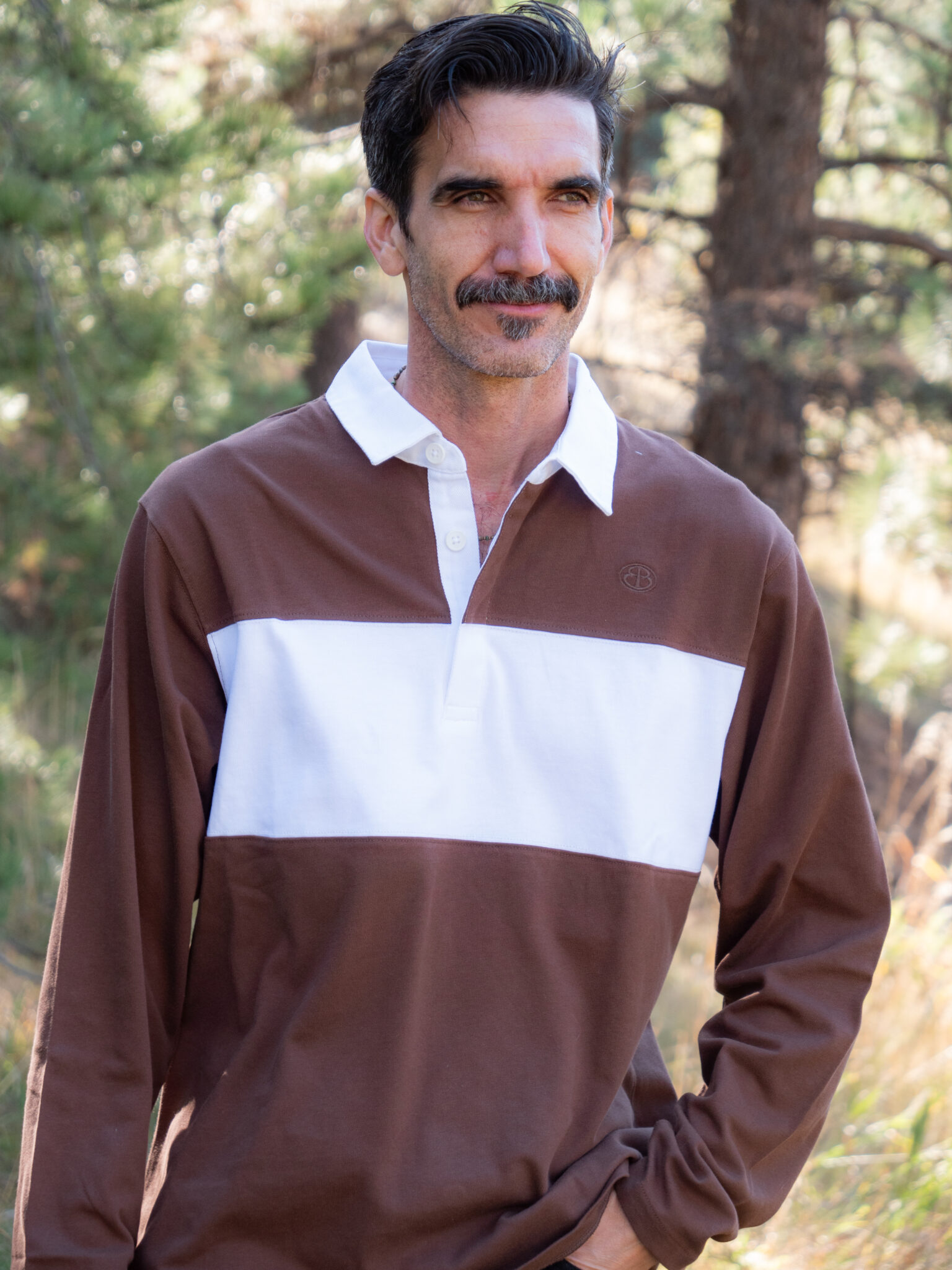 Man with dark hair and a mustache wearing a brown and white long-sleeve rugby shirt stands outdoors in a wooded area.