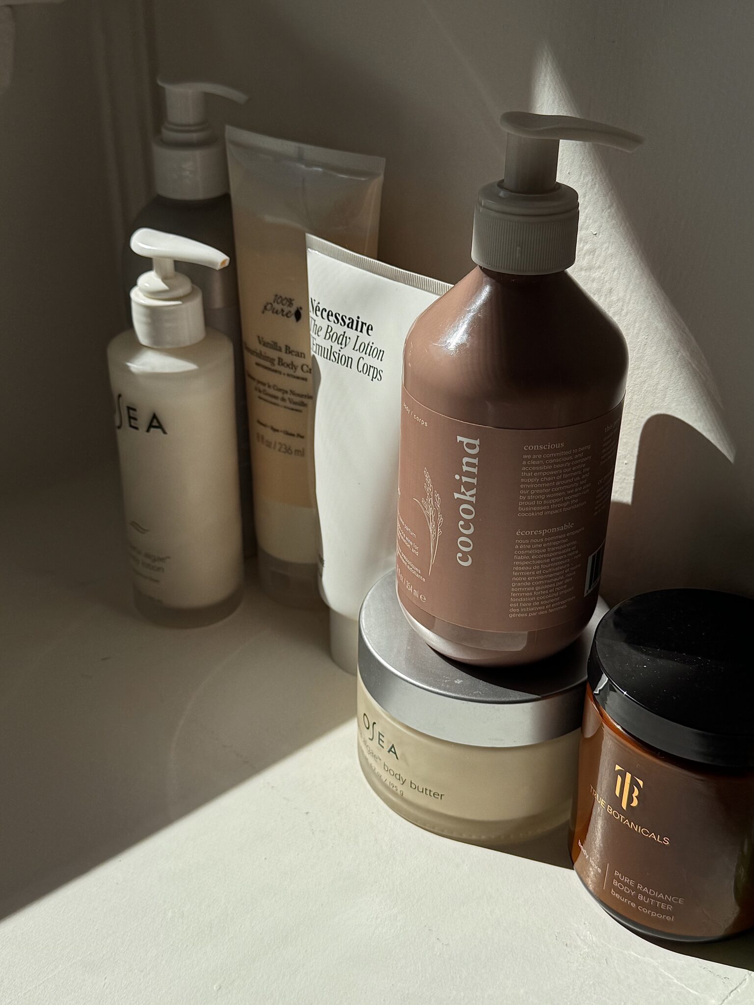 A collection of skincare and body care products, including lotions, creams, and a jar, arranged on a white surface in natural light.