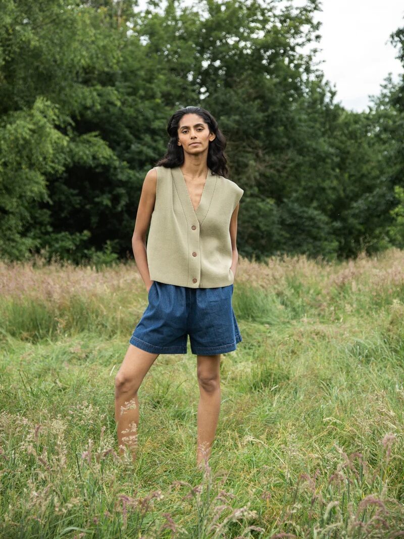 Person standing outdoors on grass, wearing a sleeveless buttoned vest and denim shorts, with green trees in the background.