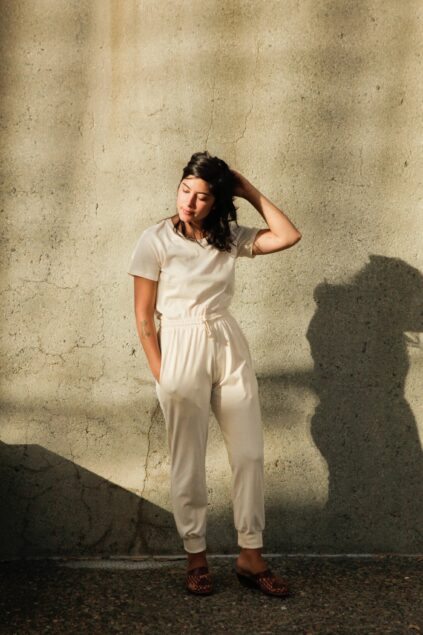 Person in a light-colored jumpsuit stands against a textured wall, hand on head, with shadow cast on the surface. Light patterns create a warm ambiance.