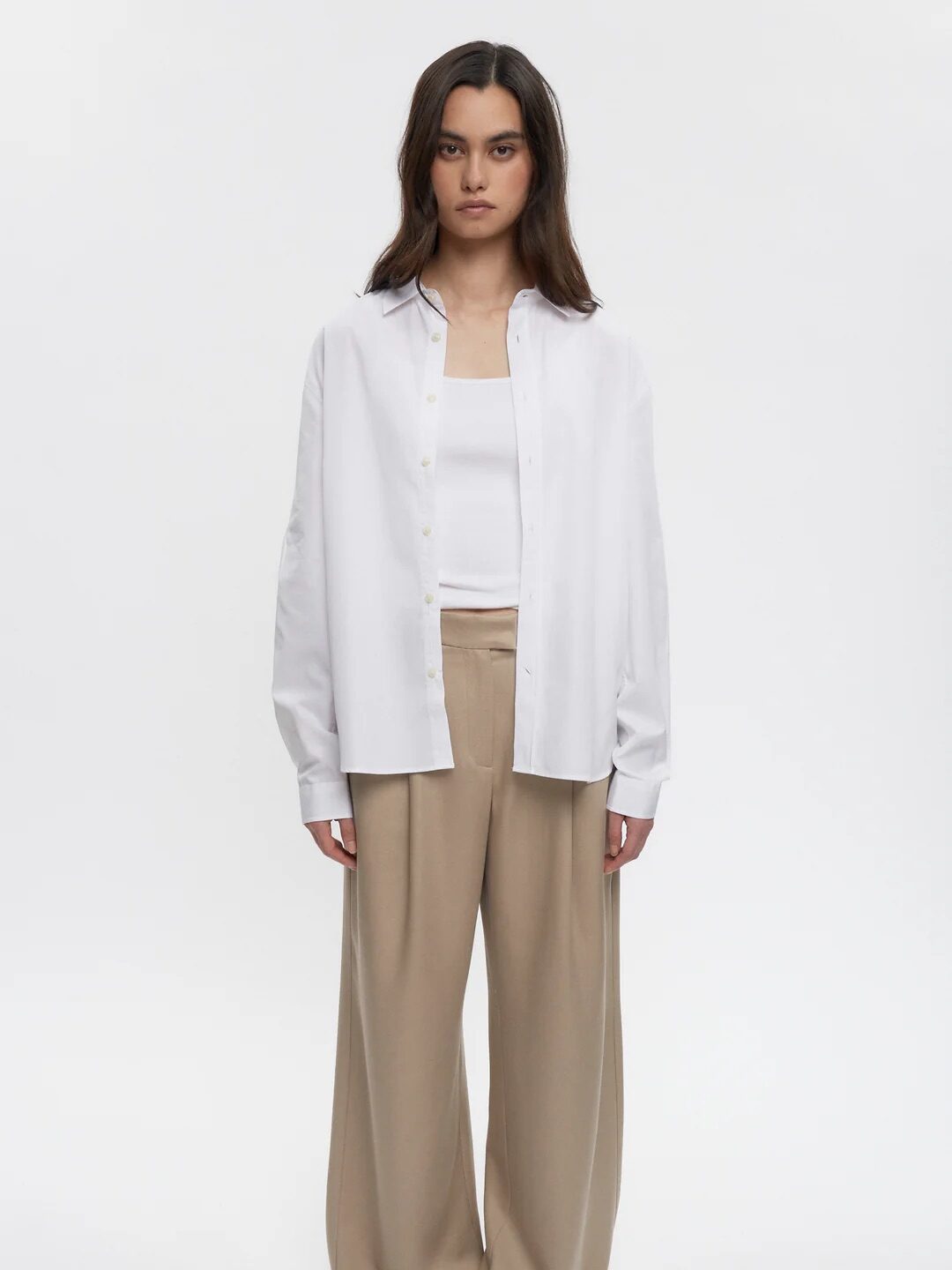 Woman standing, wearing a white shirt over a white top, beige wide-leg pants, and black sandals.