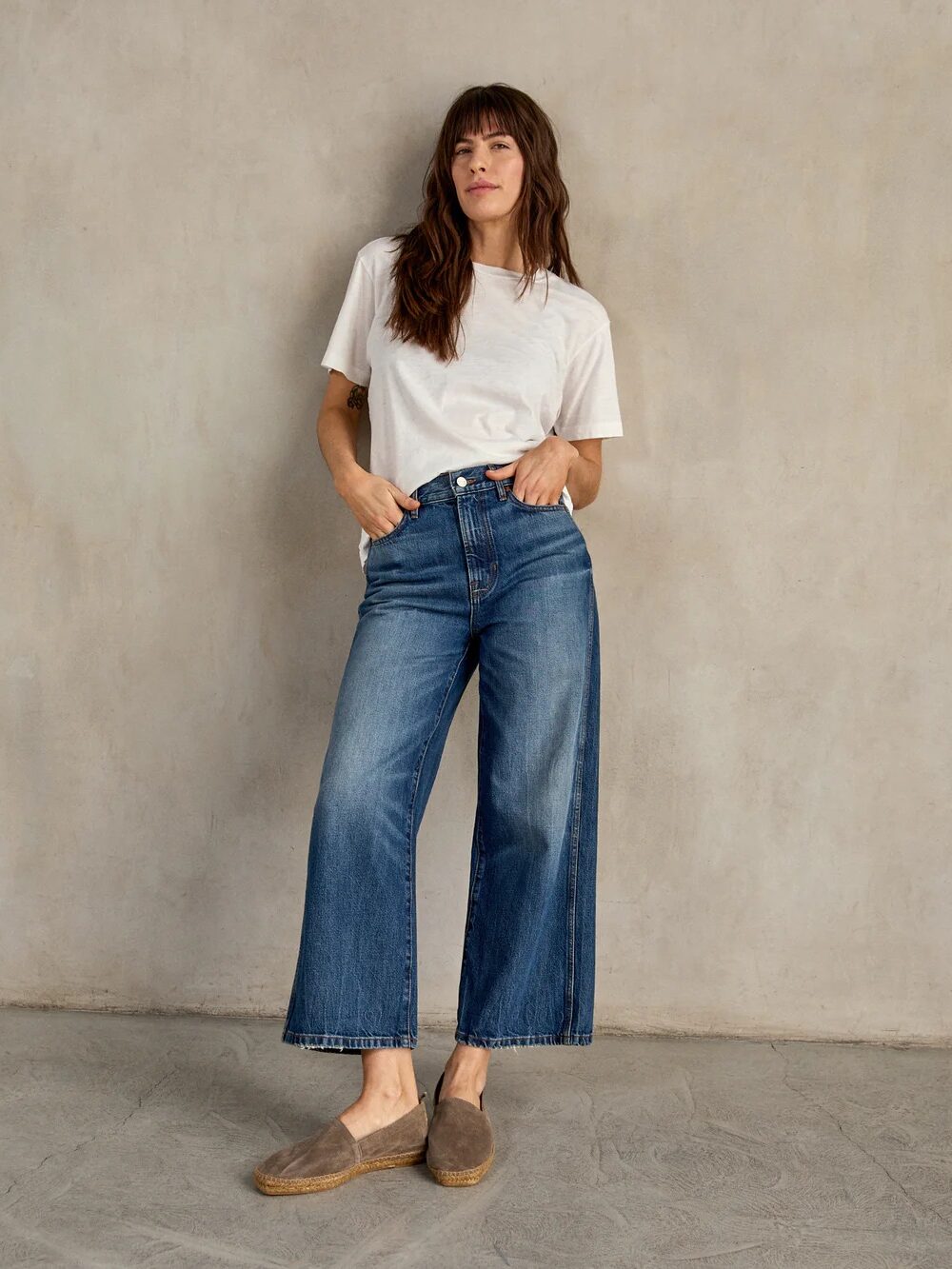 A person with long hair in a white t-shirt and wide-leg jeans stands against a plain beige background, hands in pockets, wearing brown slip-on shoes.