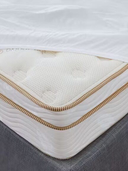 A close-up of a corner of a bed shows a mattress with a white topper partially pulled back, revealing the mattress's quilted pattern and beige piping, set on a gray bed frame over a carpet.