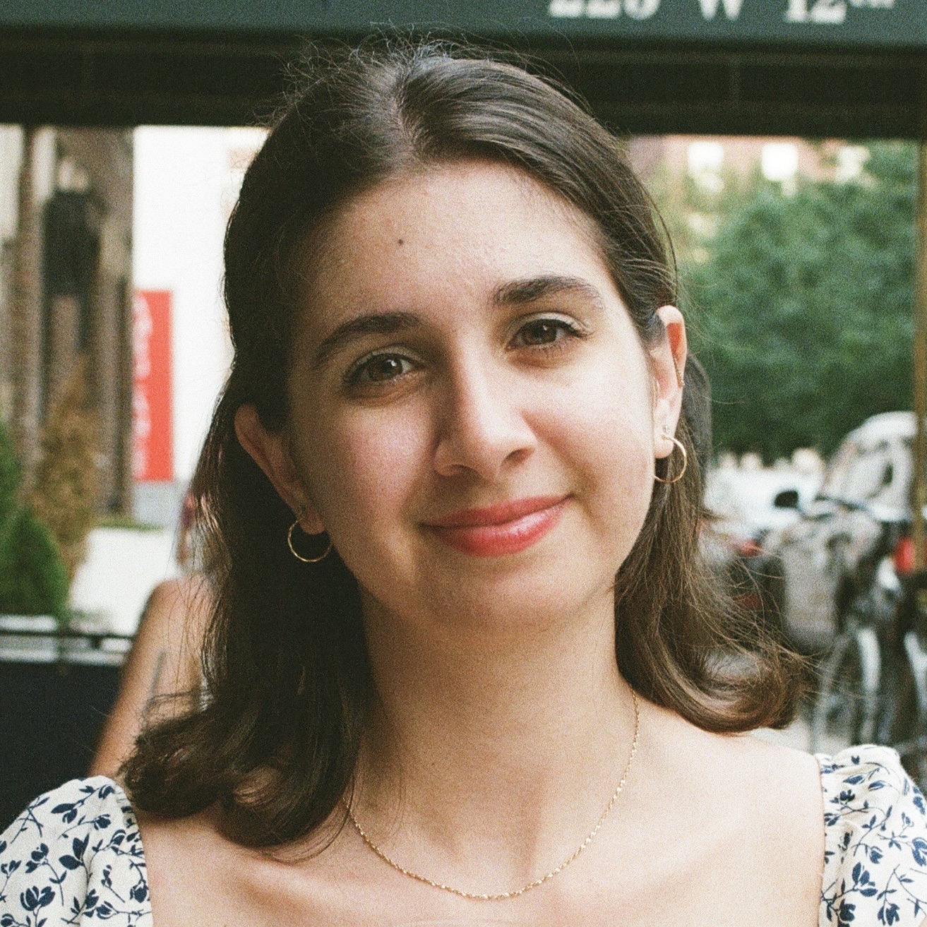 A woman with long dark hair and hoop earrings smiles at the camera. She is wearing a patterned top and standing outdoors.